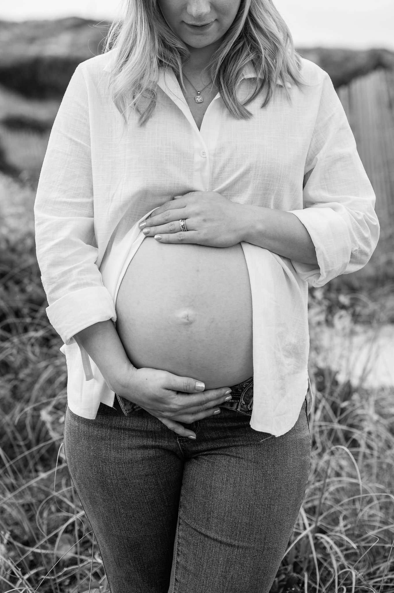 A pregnant woman standing outdoors, gently holding her belly with both hands, wearing a white button-up shirt and jeans. The photo is in black and white by Elise Souders Photography in Bend, Oregon