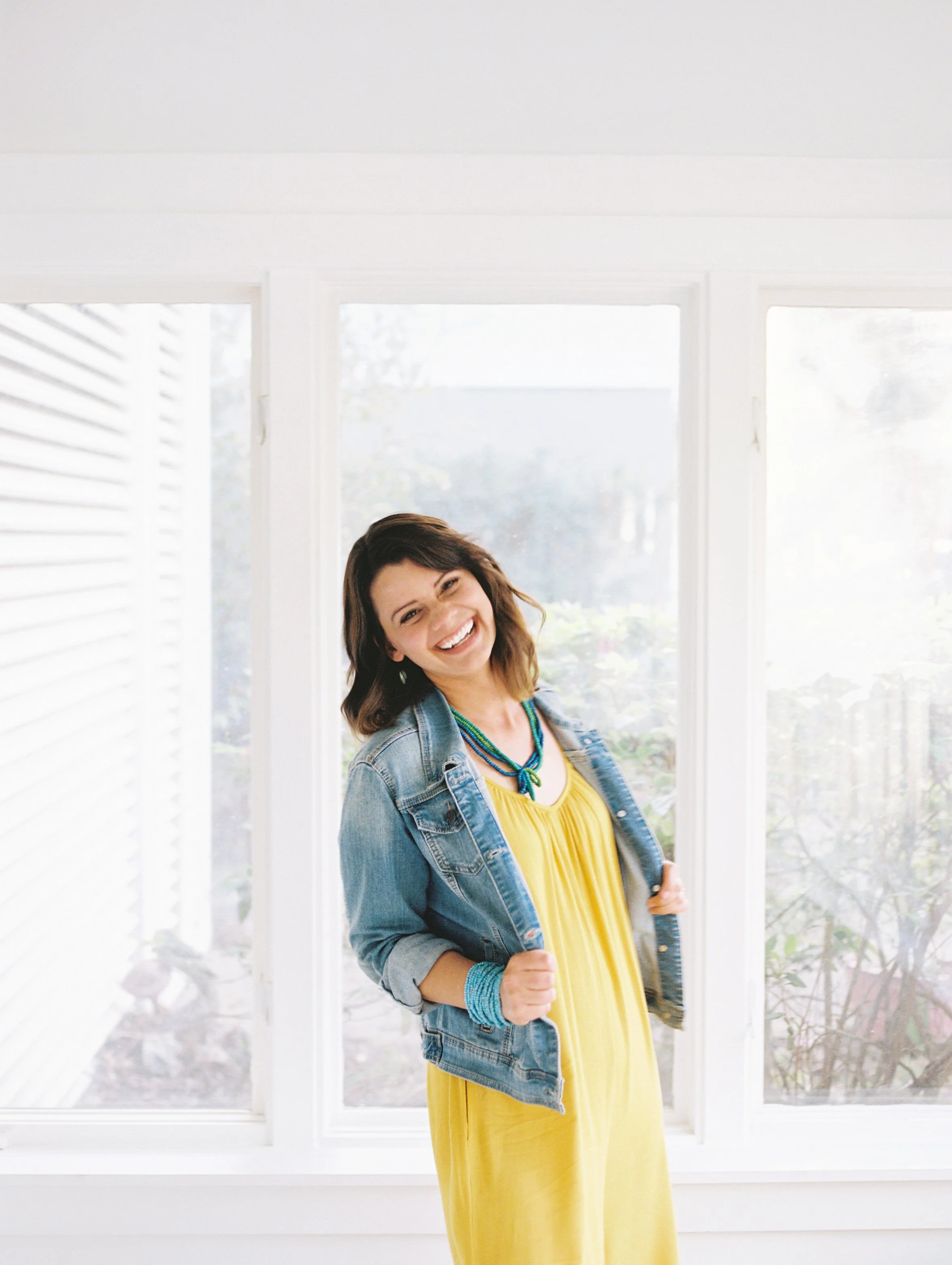 Photographer Elise Souders in a yellow dress with blue denim jacket standing by a brightly lit window, photographer Elise Souders