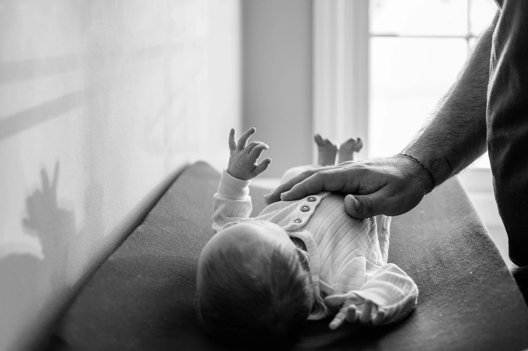 A baby lying on a changing pad while an adult gently touches the baby's arm and hand in a bright room with sunlight coming through a window in Bend lifestyle newborn photoshoot by Elise Souders