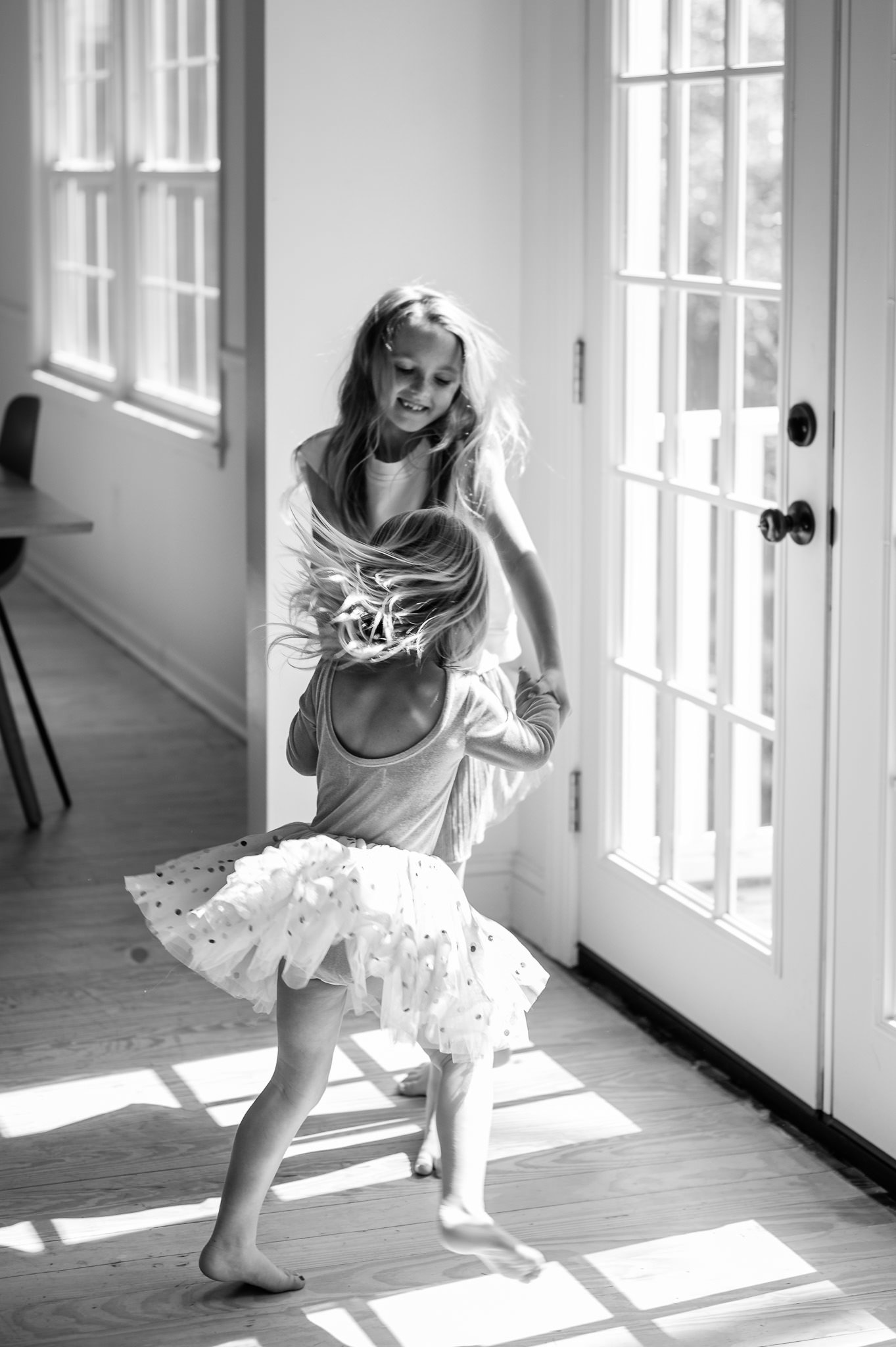 Two young girls playing and spinning near a glass door, with sunlight casting shadows on the wooden floor inside a bright room in a documentary family photo by Elise Souders Photography.
