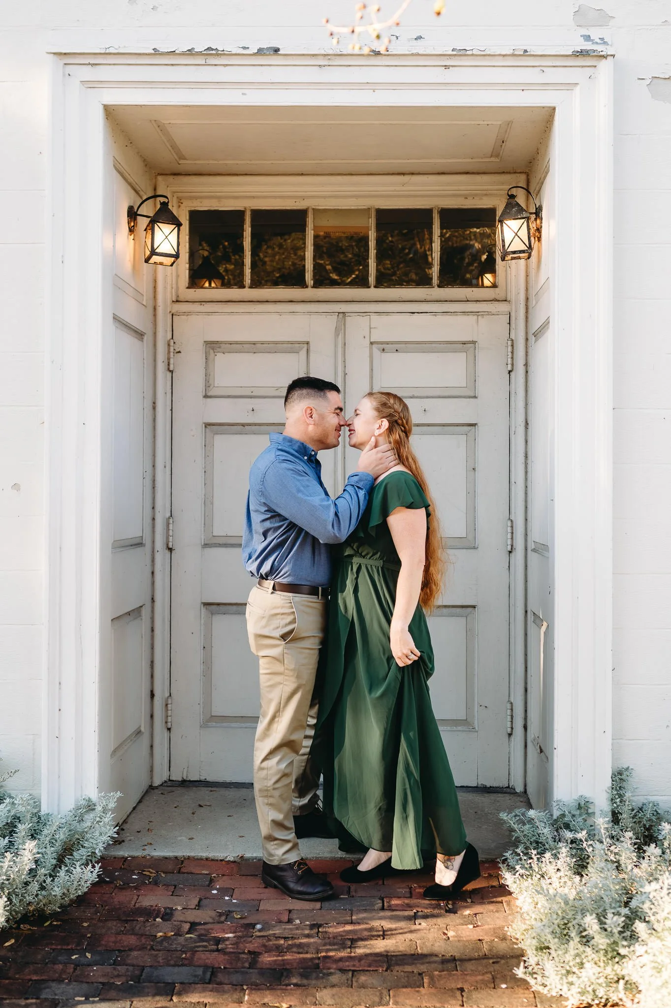 A couple standing close to each other in front of a white wooden door, smiling and touching noses, with outdoor lanterns on either side of the door illuminating them at sunset in this Bend, OR lifetyle family photo, by Elise Souders Photography.