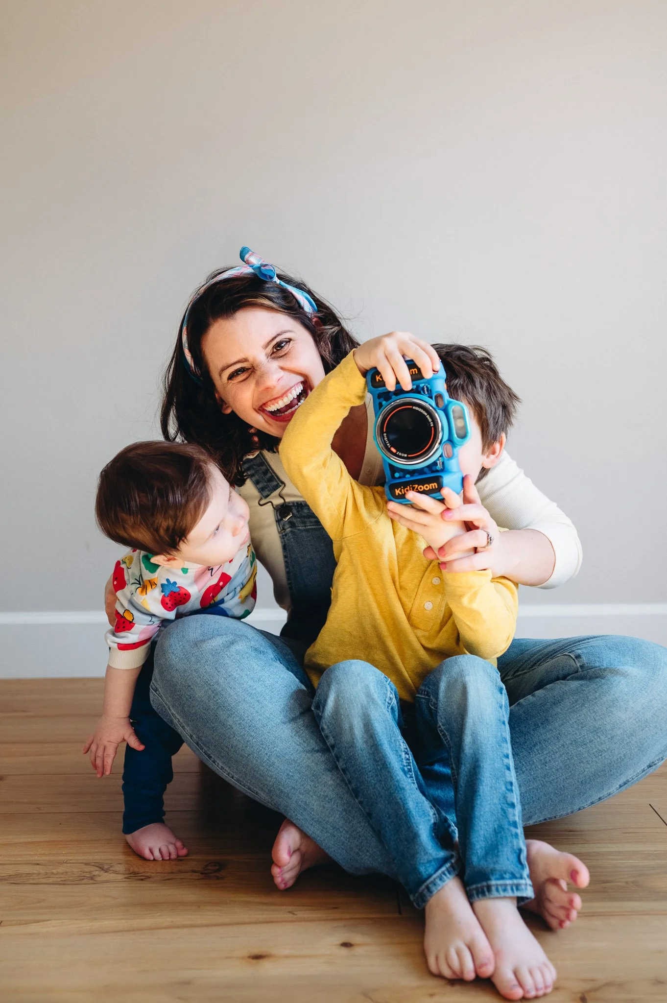 A woman and two children sitting on a wooden floor, with one child taking a photo with a brightly colored toy camera. The woman is smiling and wearing a headscarf, the younger child is wearing a patterned shirt and leaning in, and the older child is wearing a yellow shirt, holding the toy camera up to take a picture.