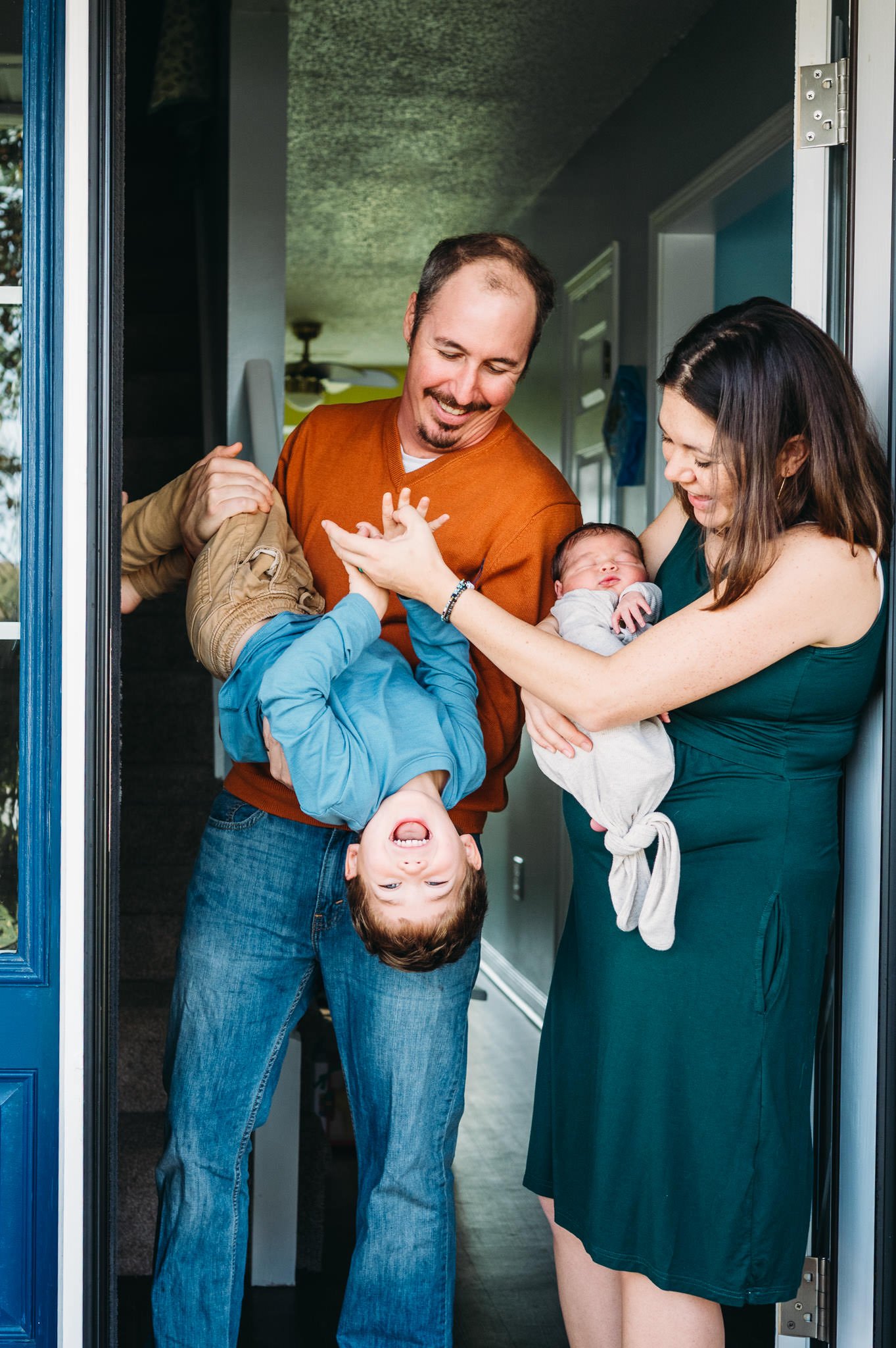 Family of four with two children, one in arms and the boy hanging upside down, standing at a front door in a fun lifestyle newborn session in-home in Bend, Oregon