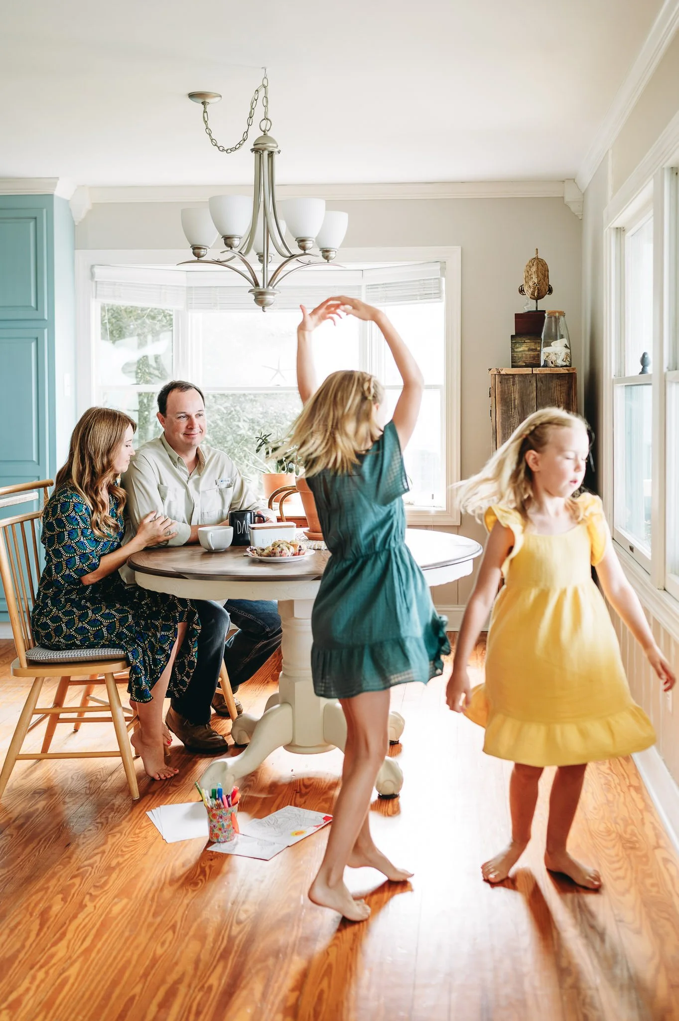 A family of four having fun in the kitchen, with two young girls dancing barefoot and adults sitting at a table in lifestyle family session at home by Elise Souders in Bend, Oregon