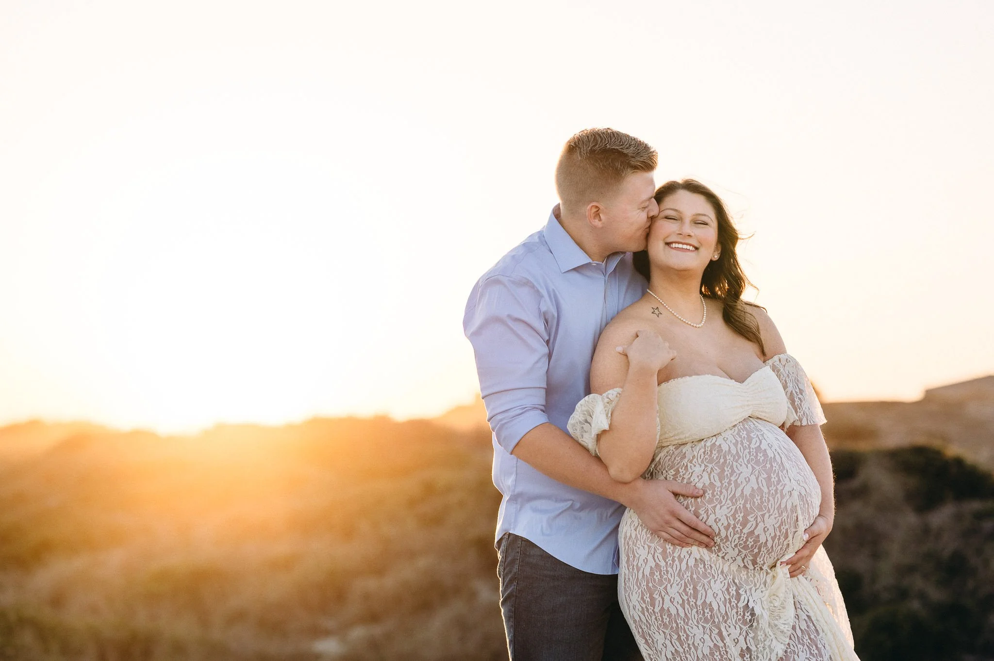 A couple, with the man kissing the woman on the cheek, stands outdoors during sunset. The woman is pregnant and smiling, wearing a lace off-shoulder dress and a pearl necklace. Golden hour maternity photo of expecting couple in Central Oregon 