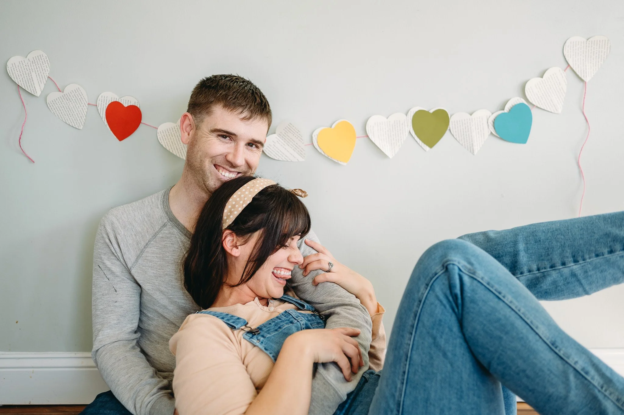 A happy couple sitting on the floor, with the woman laughing and the man smiling behind her, in a room decorated with colorful paper hearts hanging as a garland.