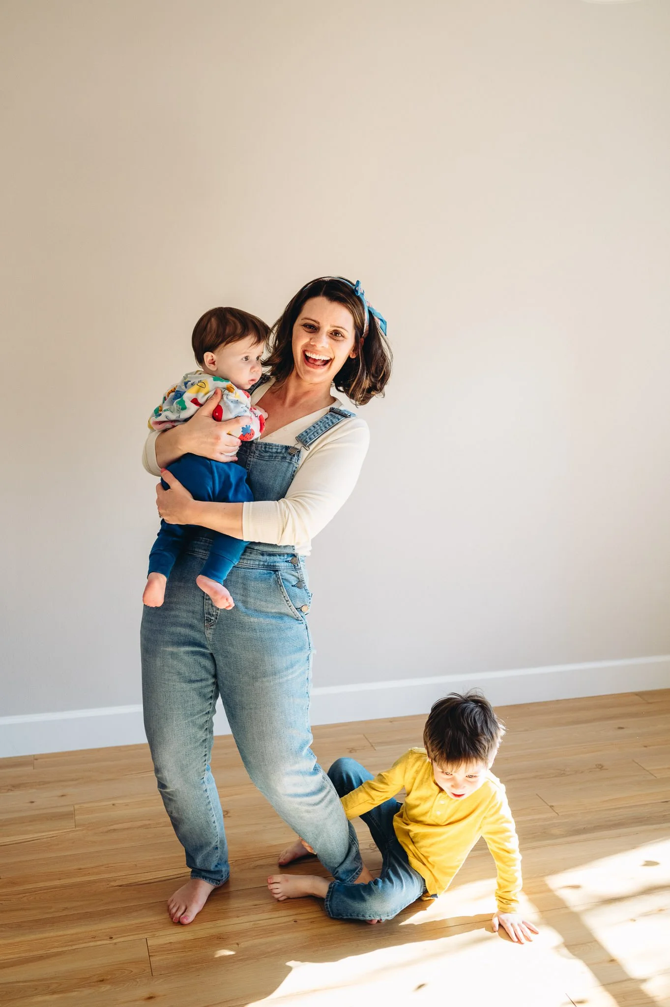 A woman holding a baby in her arms while standing on a wooden floor, with a young boy sitting on the floor nearby. The woman is smiling and wearing denim overalls and a cream shirt, and the boy is wearing a yellow shirt and jeans, appearing to be playing or crawling. The scene is bright with natural light.