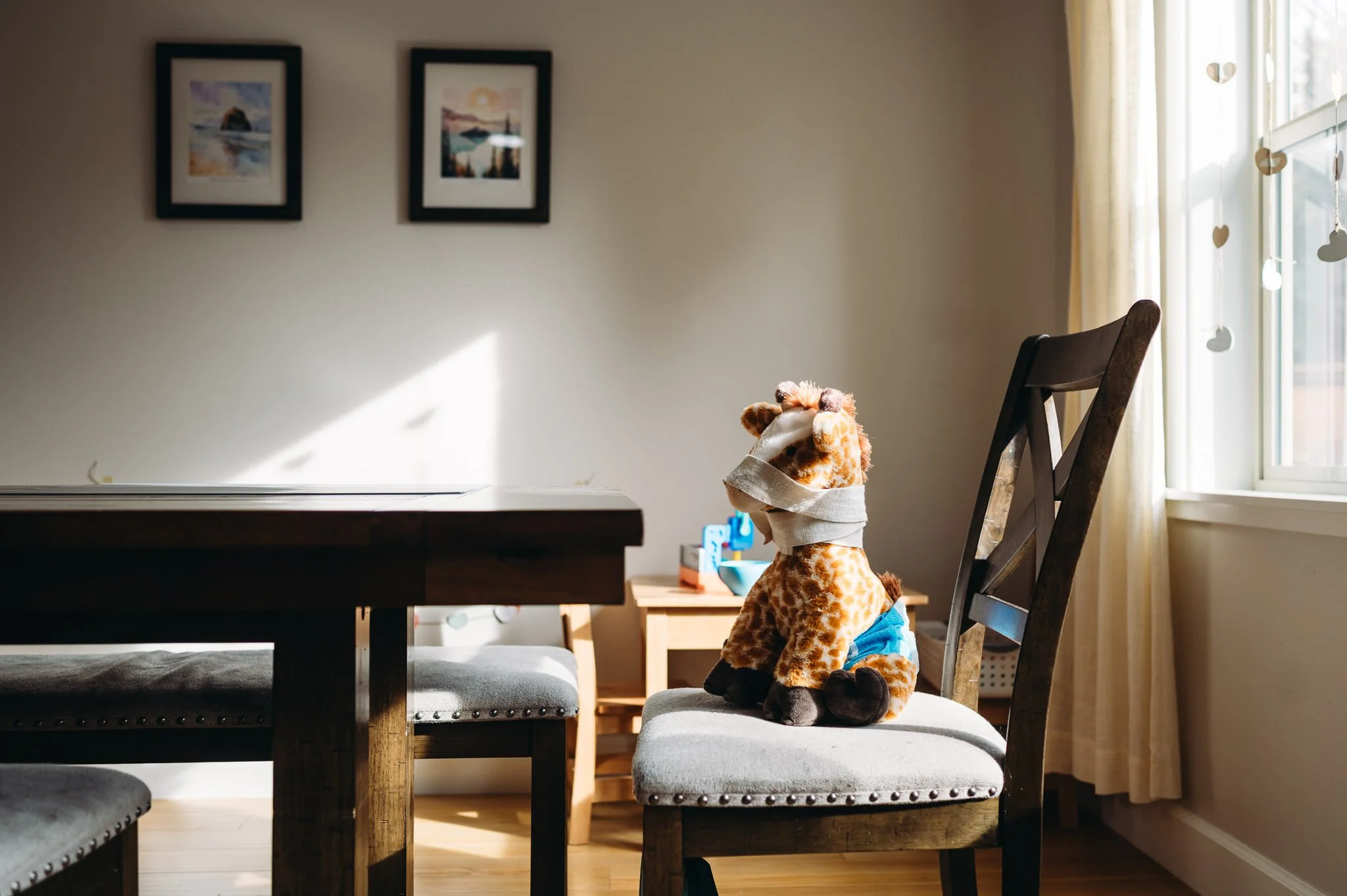 A stuffed giraffe toy sitting on a dining chair in a bright dining area wearing a pull-up and with a pretend bandage wrapped around it in a documentary family photo by Elise Souders Photography