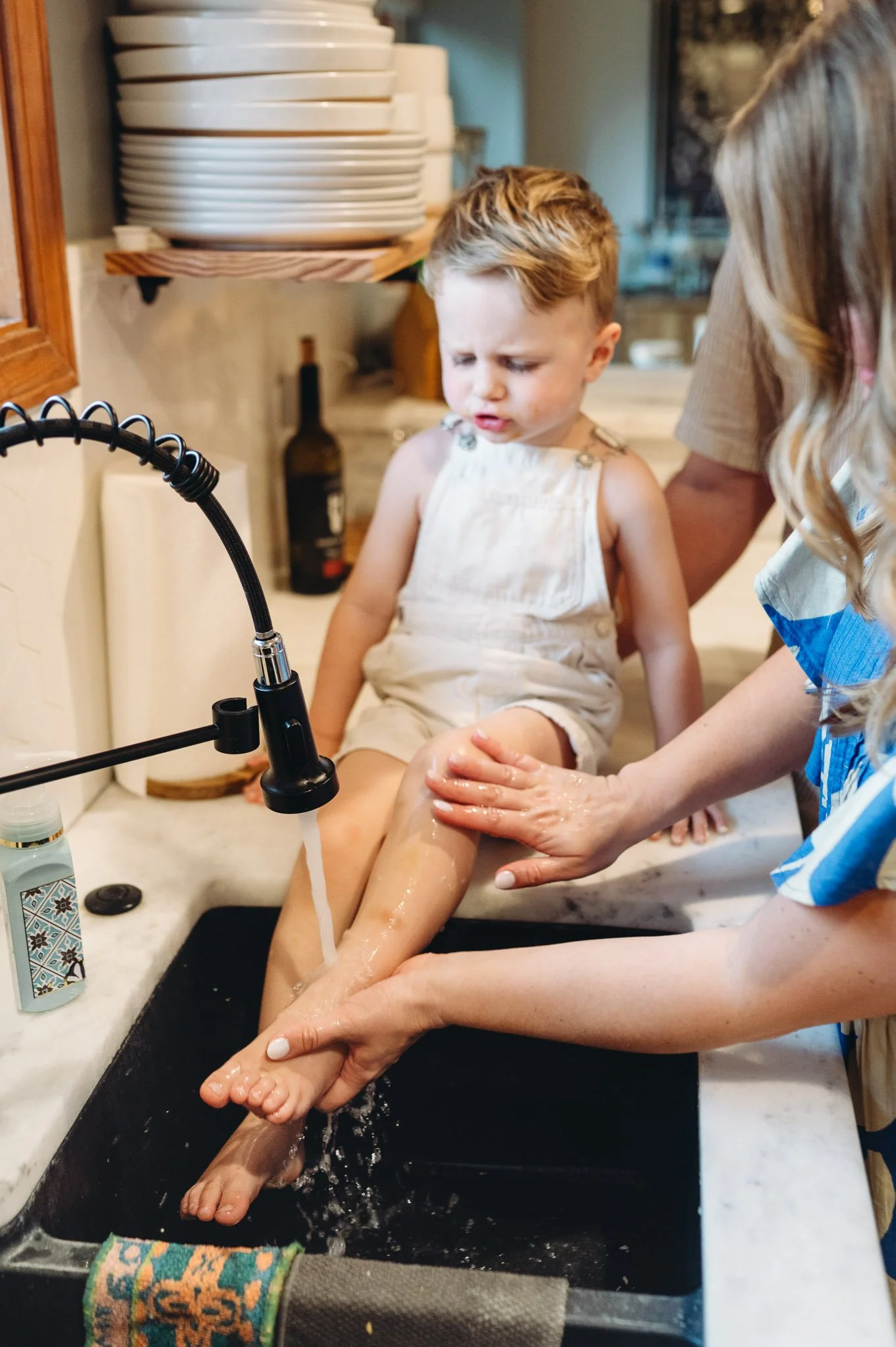 A young boy sitting on a kitchen counter getting his legs washed by a woman, while another woman helps, in a home kitchen.