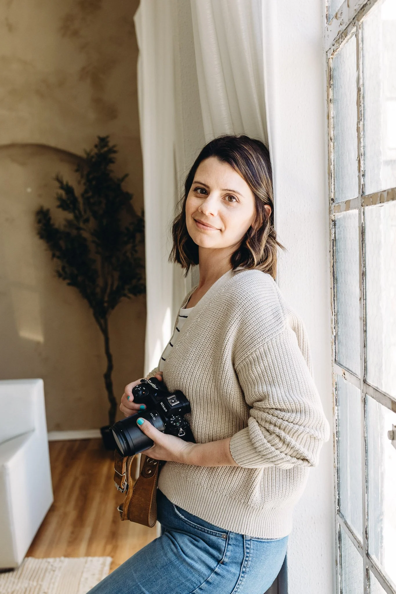 Elise Souders, Bend family photogapher, stands near a window with white curtains in a room with wooden floors. She is holding a black Nikon camera in her right hand and looking at the camera with a slight smile.