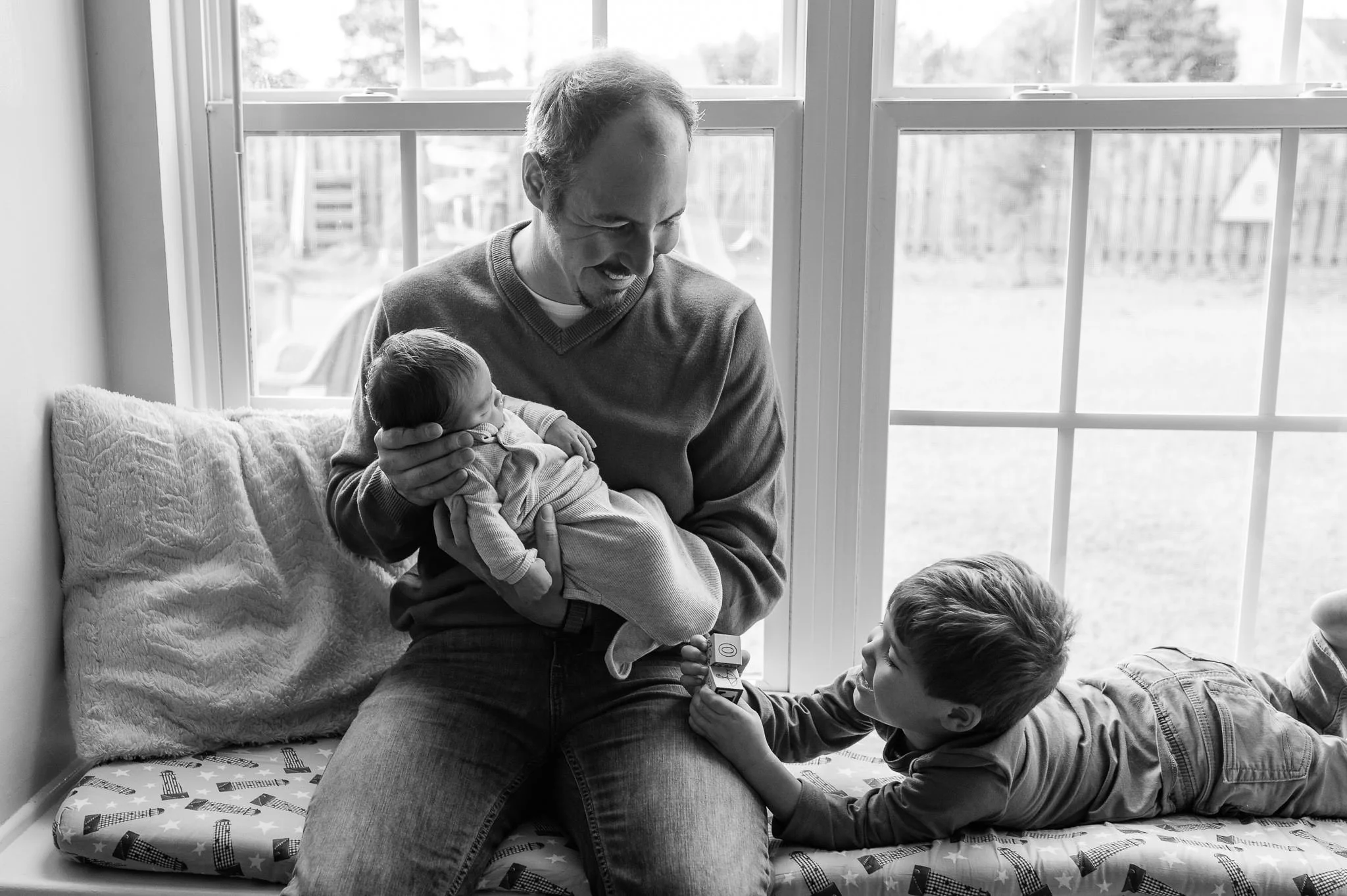 A man holding a baby in his arms, smiling and looking at a young boy lying on a bench and reaching for building blocks, near a window with a view of a backyard in-home lifestyle newborn photoshoot by Elise Souders