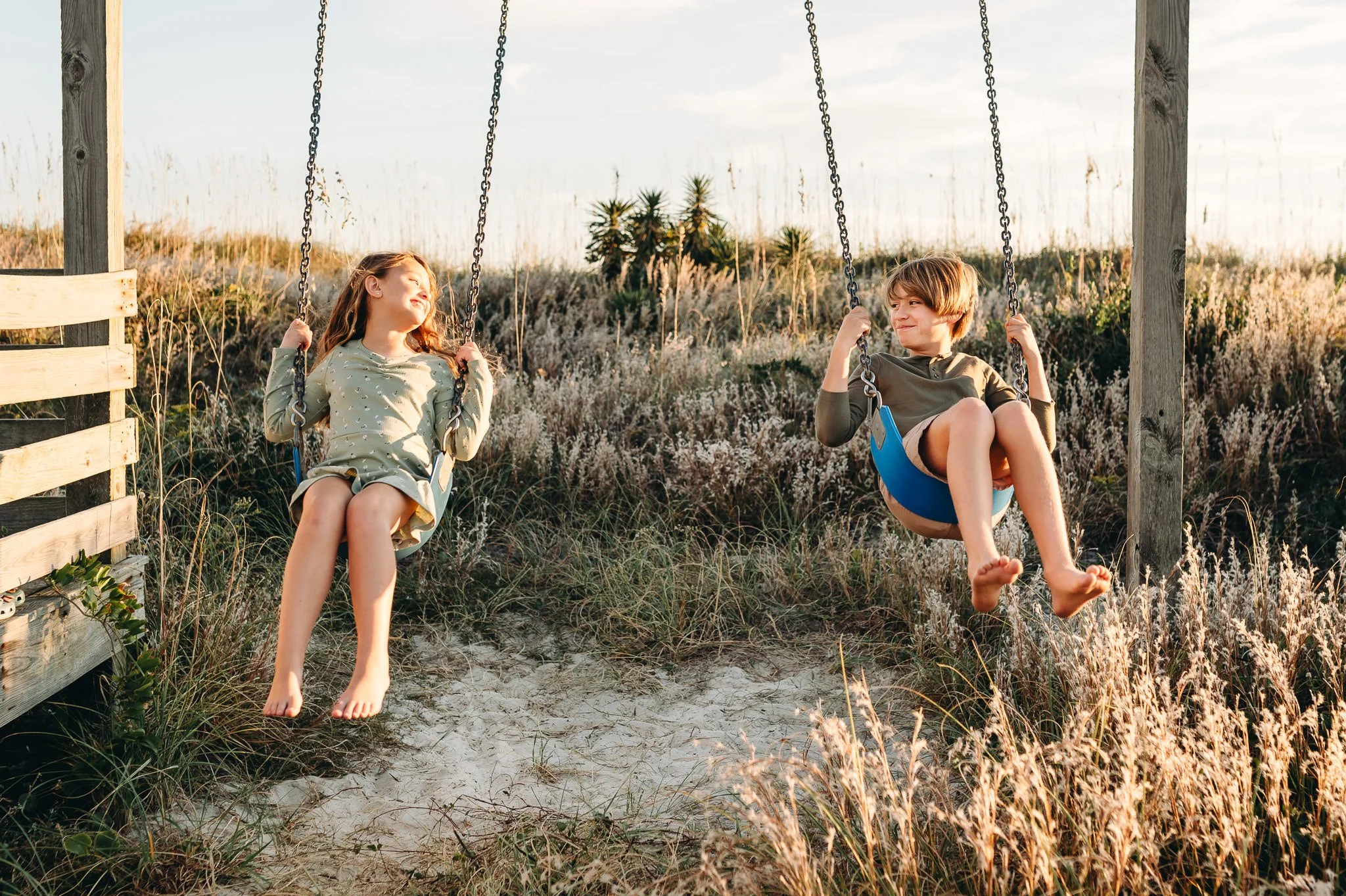 Siblings smile at each other while they swing side by side in golden sunset lighting, Bend lifestyle family photography.
