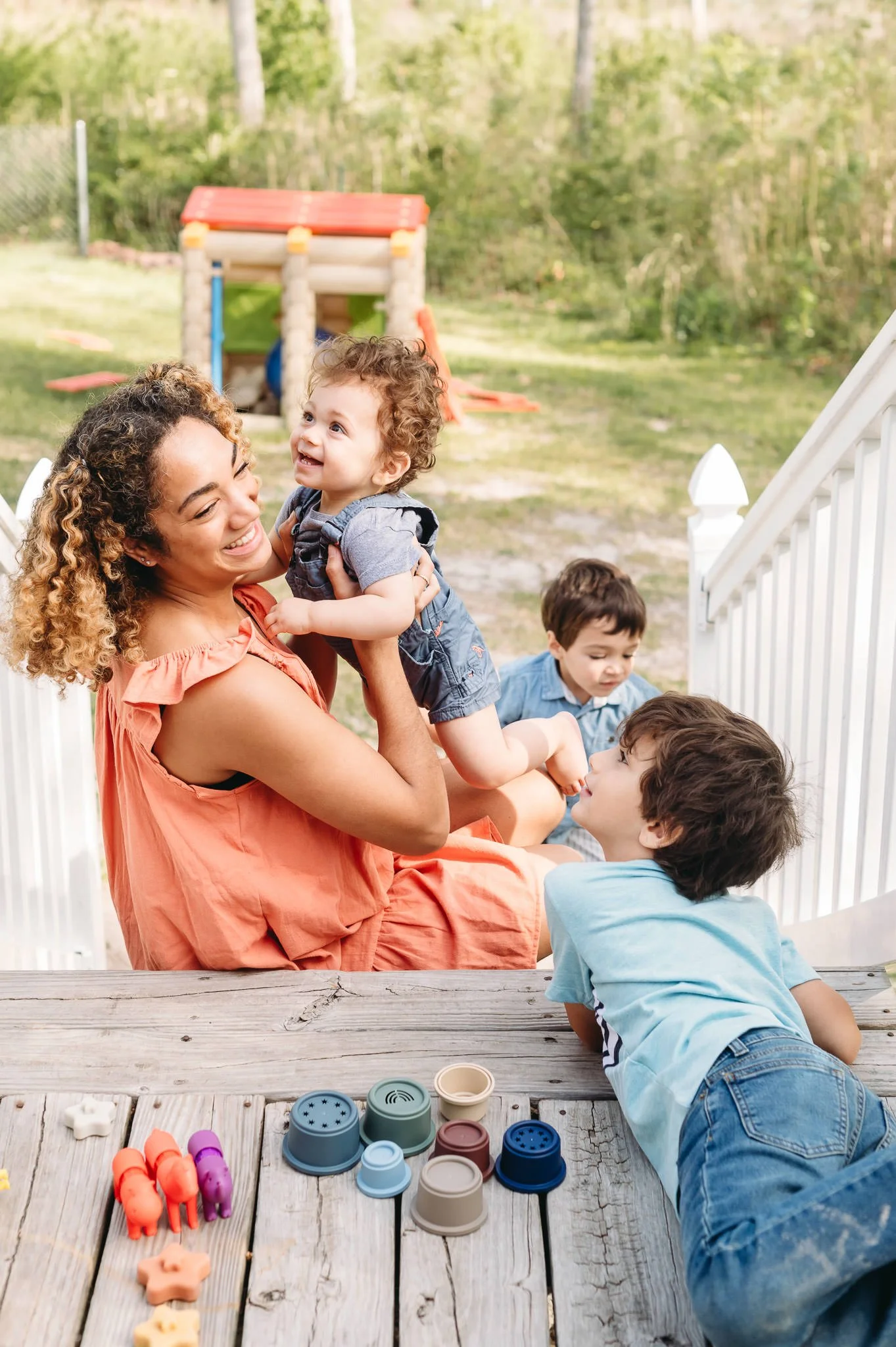 A woman and three children playing outdoors on a deck with a white railing. The woman holds a curly-haired toddler and smiles at her. Two boys sit on the deck near her, one lying on his stomach and the other sitting beside her. There are colorful toys and plant pots on the wooden surface of the deck, with a grassy yard and a playhouse in the background.