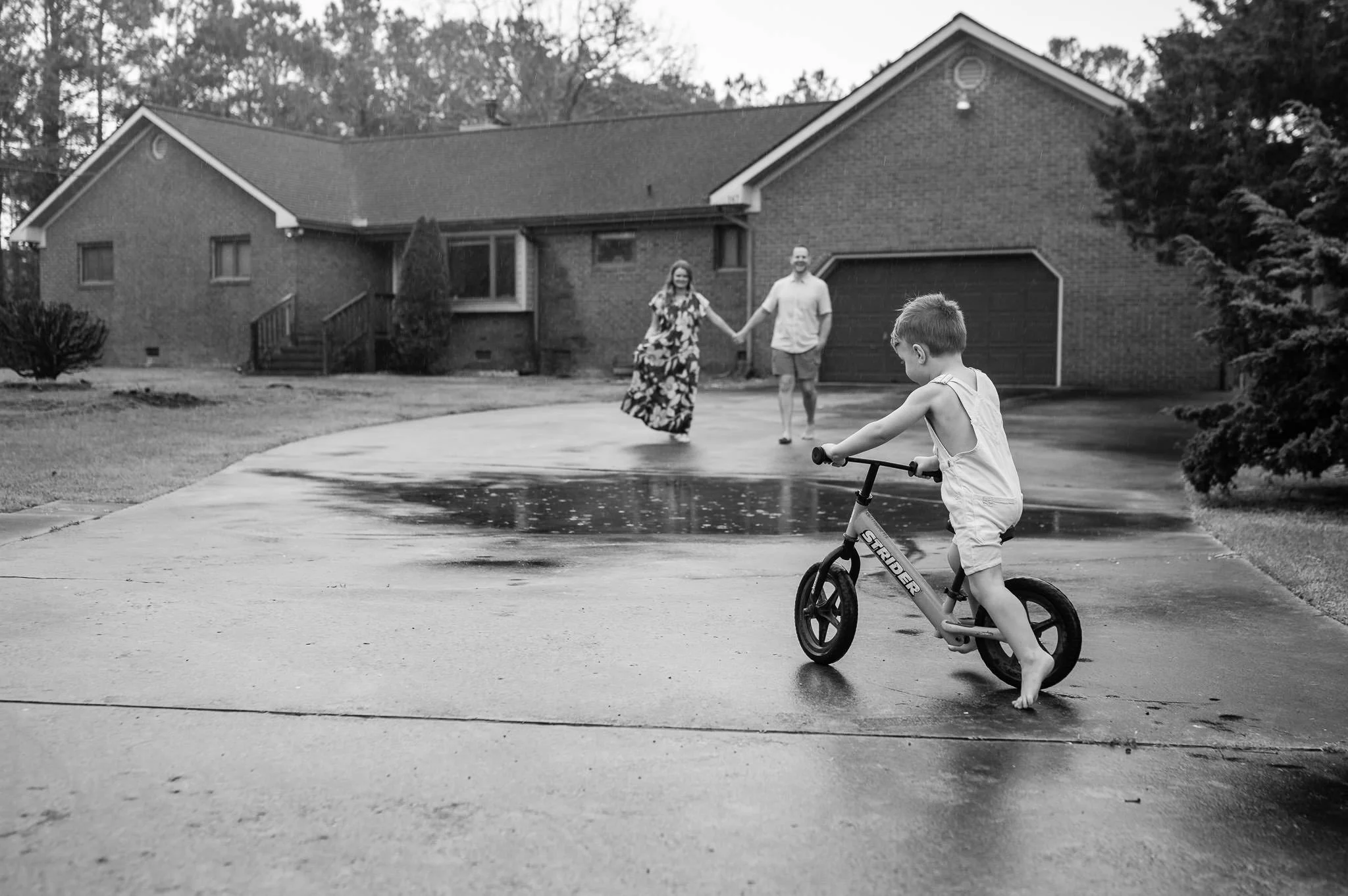 A young boy on his balance bike in the driveway in front of his home with his parents holding hands in the background, lifestyle family photographer Central Oregon. 