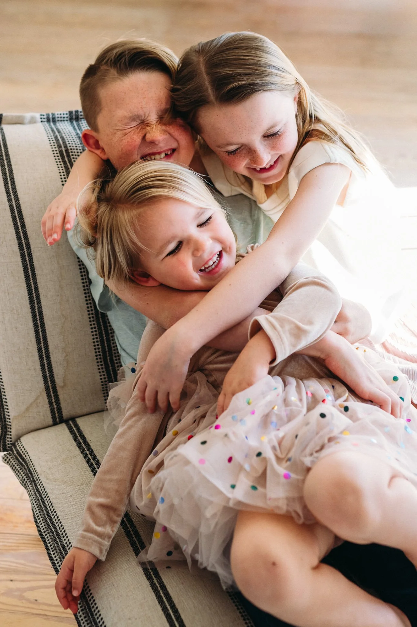 Four children laughing and hugging on a sofa in a brightly lit room in-home photography in Bend, Oregon.
