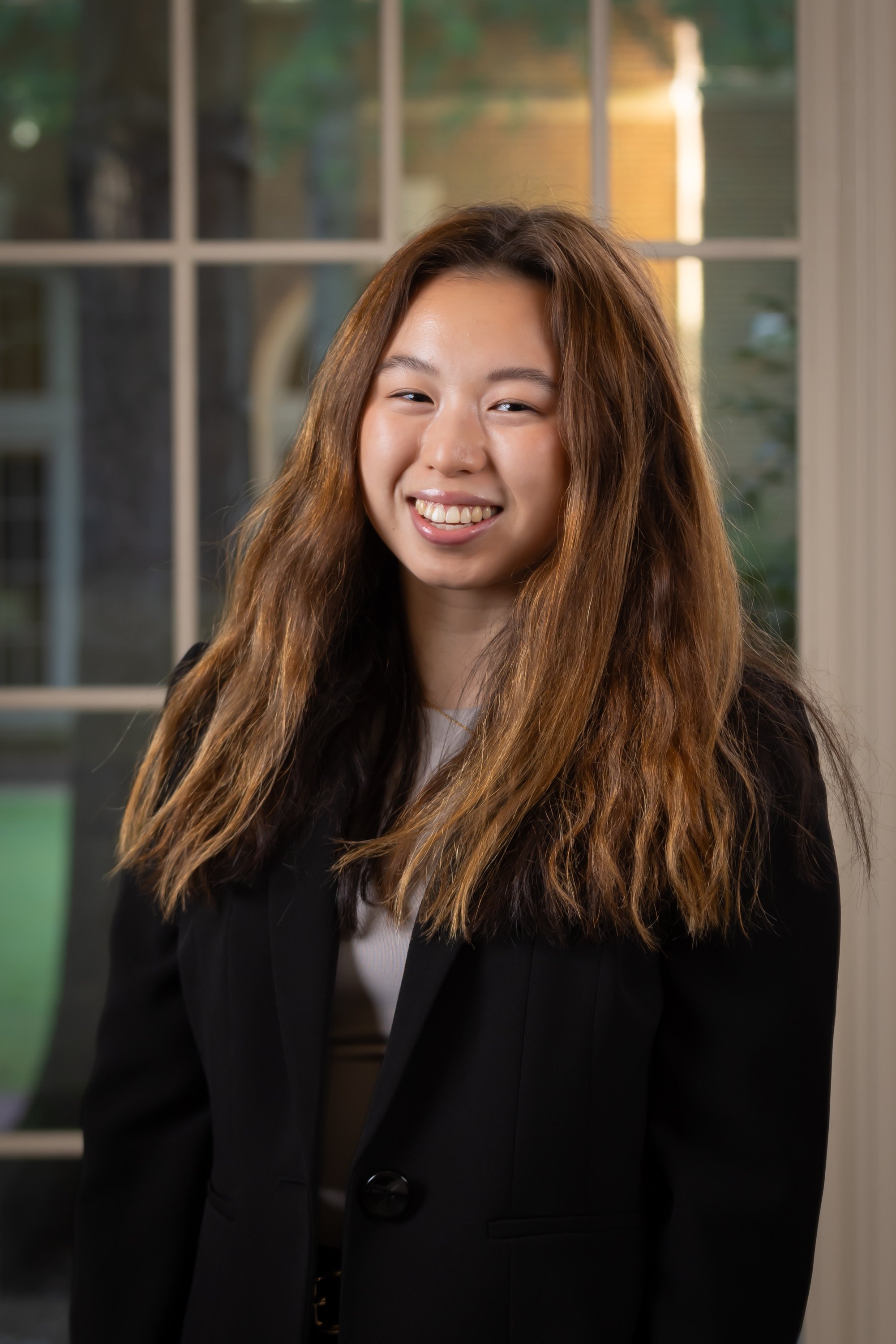 A young woman with long, wavy brown hair smiling in front of a large window with a blurred outdoor background.