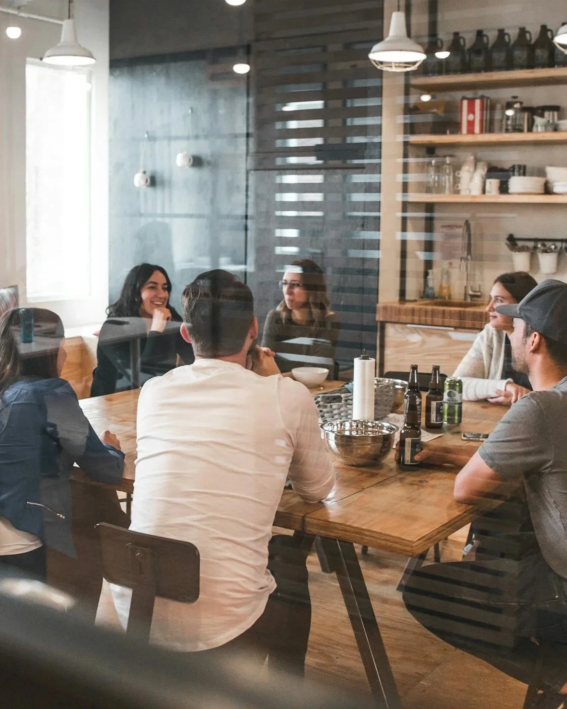A group of six young adults sitting around a wooden table in a modern café or restaurant, engaged in conversation and laughter, with drinks and snack bowls on the table, seen through a glass window.