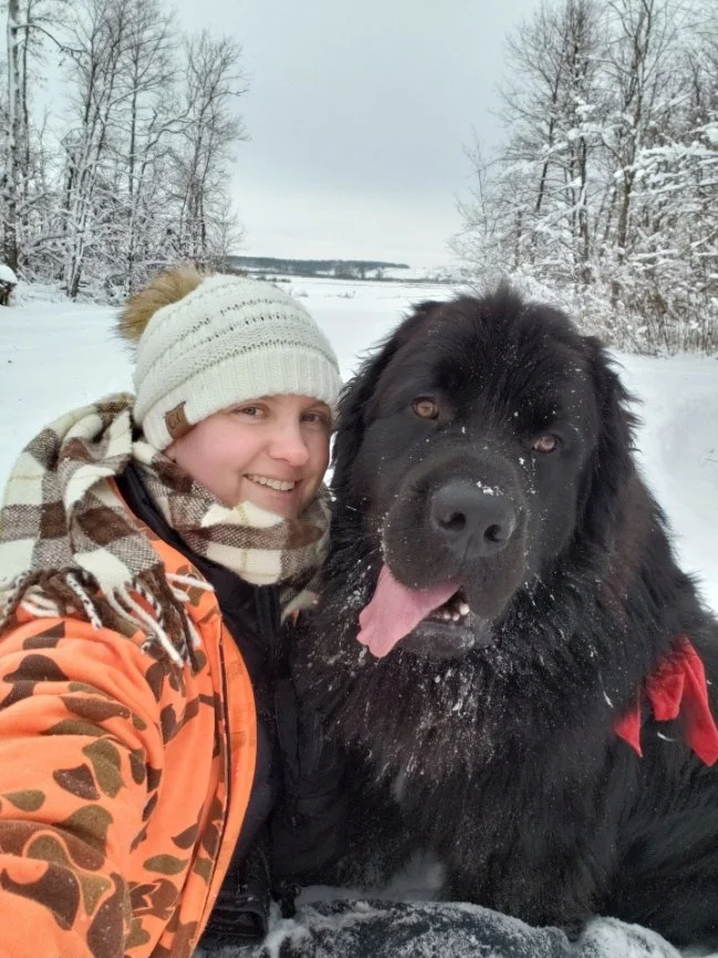 A woman in winter clothing and a black Newfoundlander dog outdoors in a snowy landscape with trees and a frozen body of water in the background.