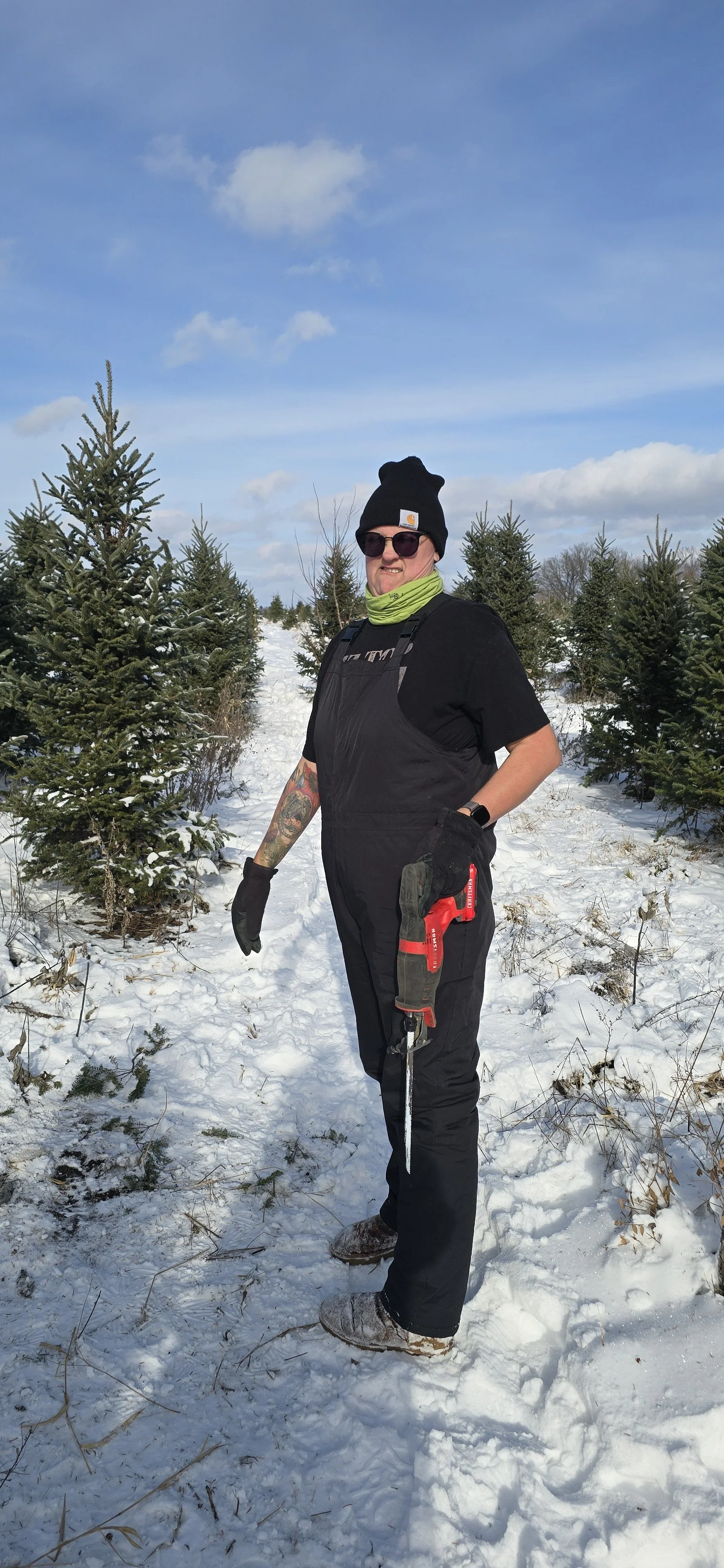Person standing in snow-covered area with small Christmas trees, wearing a black beanie, sunglasses, green neck gaiter, black gloves, black overalls, and holding a red chainsaw.
