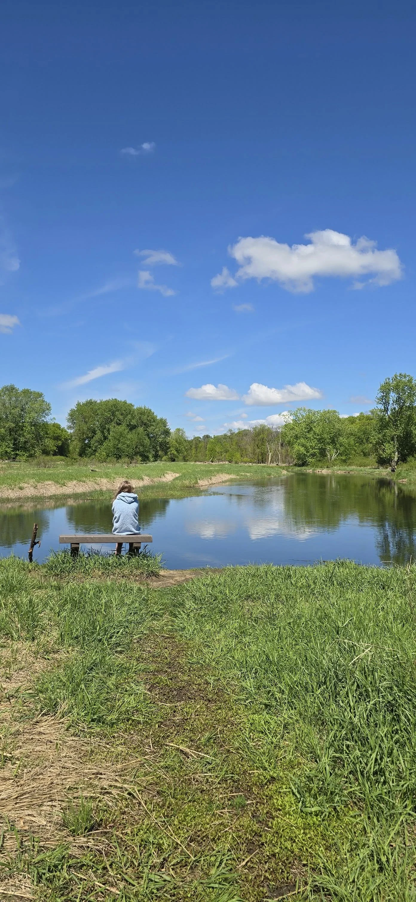 A person in a gray hoodie sitting alone on a wooden bench by a calm river, with green trees and a blue sky with scattered clouds reflected in the water.