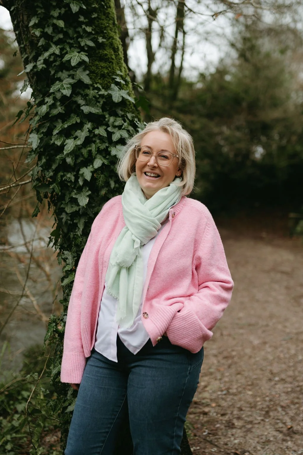 A woman with glasses and blonde hair, wearing a pink cardigan, white shirt, and a light green scarf, smiling and standing outdoors next to a tree covered in ivy.