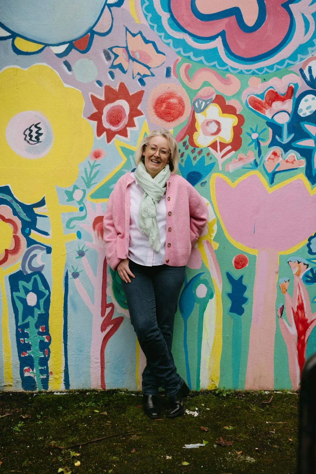 A woman standing in front of a colorful floral mural, smiling, wearing glasses, a pink cardigan, white shirt, blue jeans, and black boots.