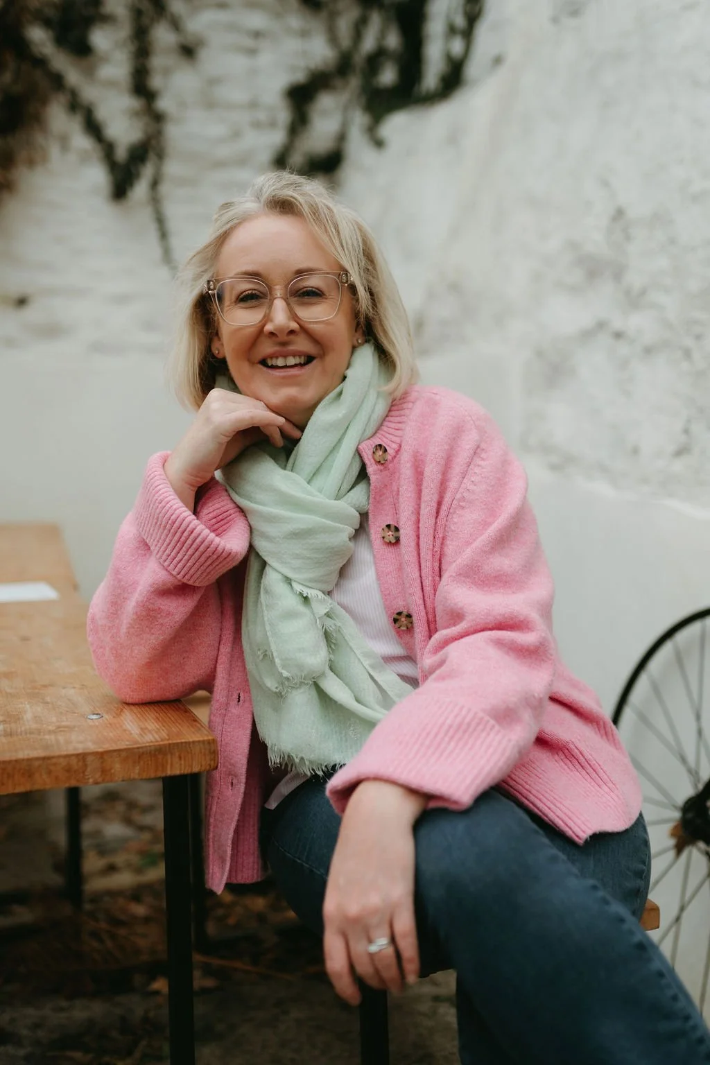 A woman with blonde hair, glasses, wearing a pink cardigan, light green scarf, and a white top, sitting at a wooden table indoors, smiling at the camera.