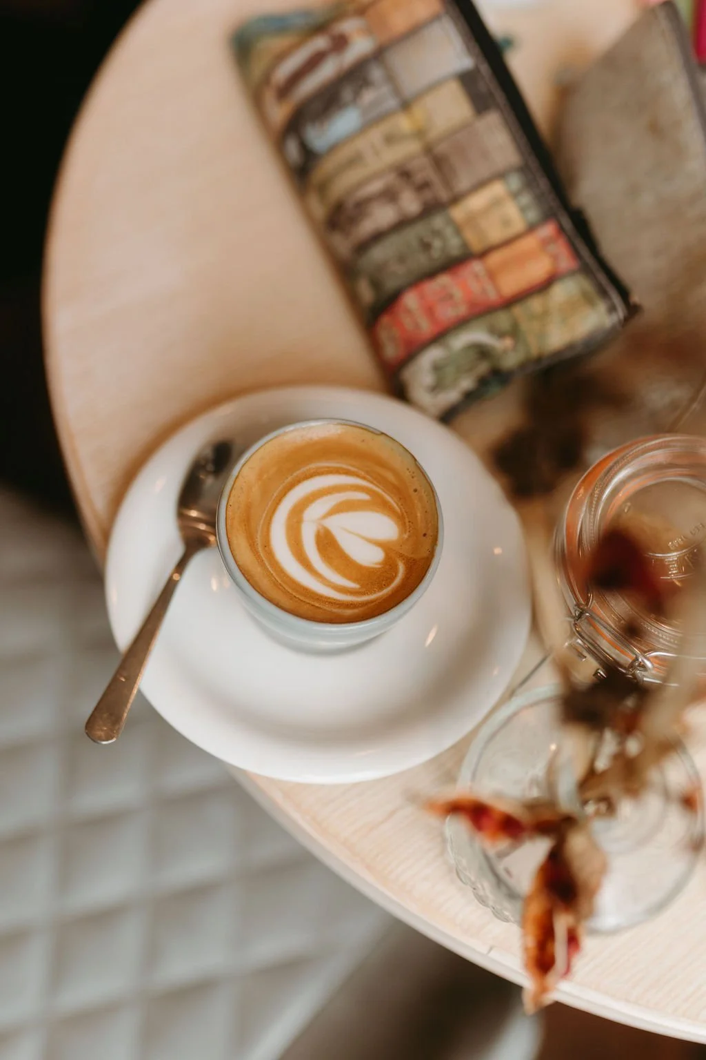 A cup of coffee with latte art on top, placed on a saucer with a spoon, on a wooden table with a can of sardines and a jar of dessert with a spoon in it.