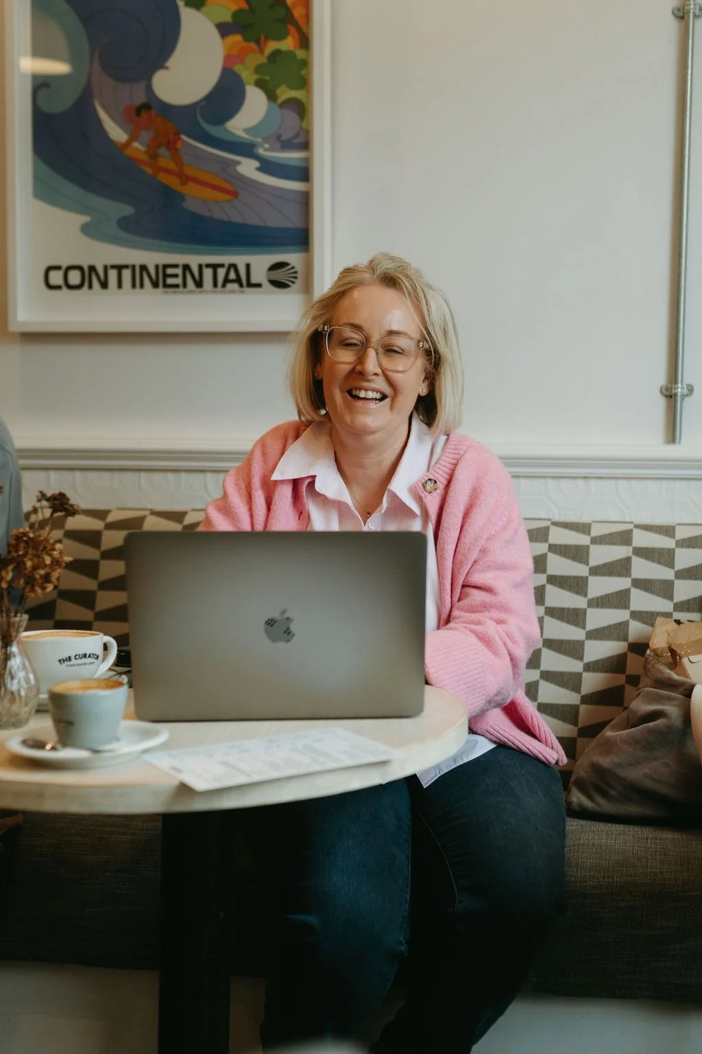 A woman with glasses and blonde hair, wearing a pink cardigan and white shirt, sitting at a table with a laptop, smiling and laughing in a cozy cafe.