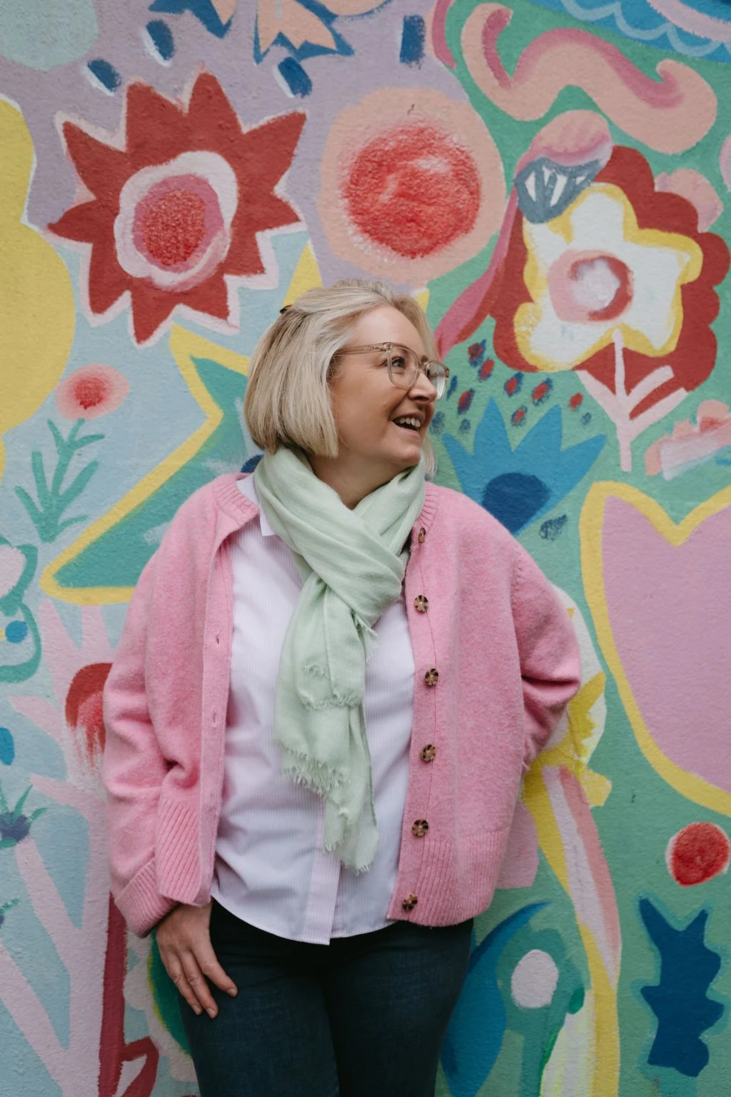 A woman with short blonde hair and glasses smiling, standing in front of a colorful mural with flowers and abstract shapes, wearing a pink cardigan, white shirt, and light green scarf.