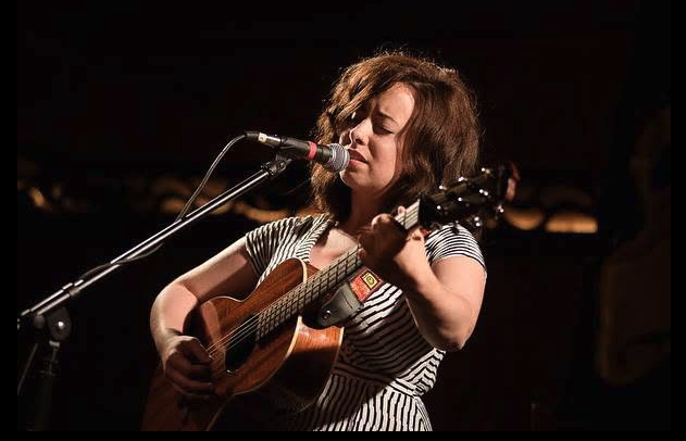 Lauren Pratt, musician, playing guitar and singing into microphone with eyes closed in darkened room.