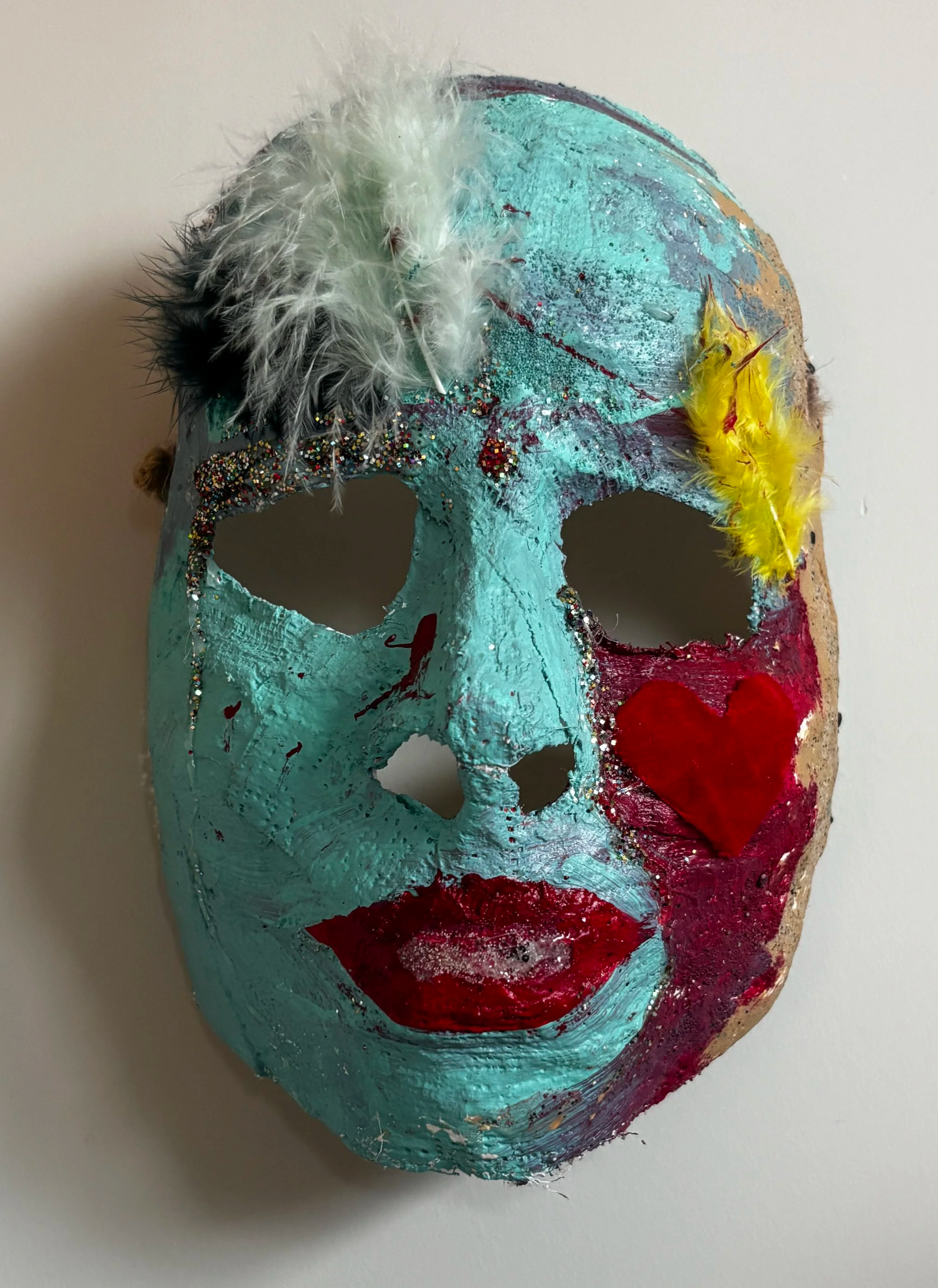 A plaster mold of a face with teal paint, red lips, and feathers applied.