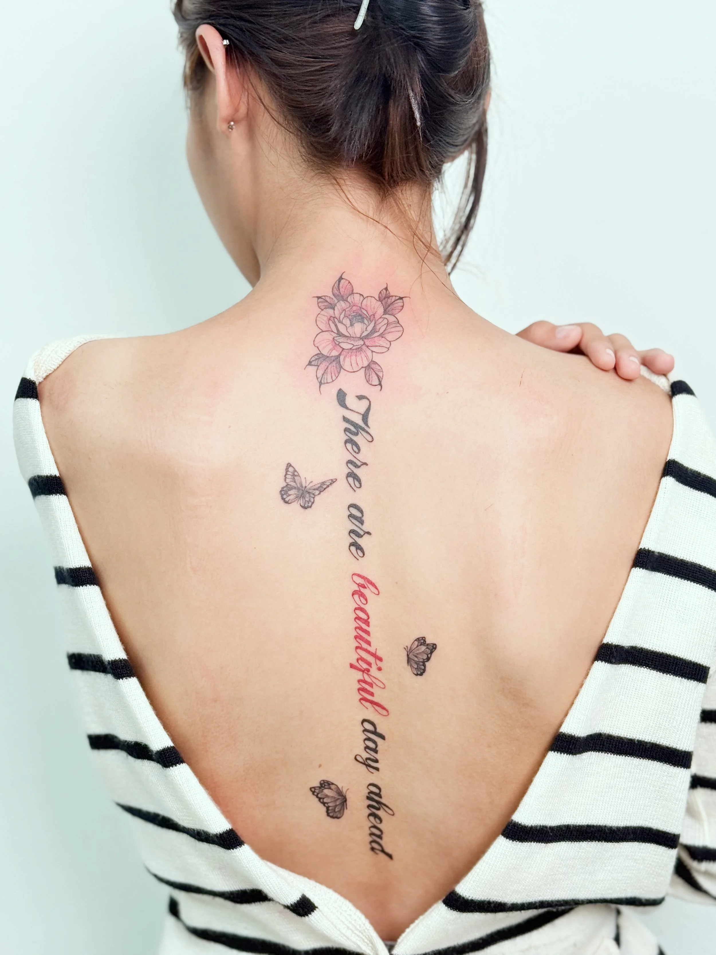 รอยสักดอกไม้และตัวอักษรสไตล์ Fine Line บริเวณแผ่นหลังจาก Let’s Tatt Studio / Fine line floral and lettering tattoo by Let’s Tatt Studio Chiang Mai ดอกไม้และลายเส้นที่เรียงตัวตามแนวกระดูกสันหลัง สื่อถึงการเติบโต ความอ่อนโยน และตัวตนที่ค่อย ๆ เบ่งบานตา