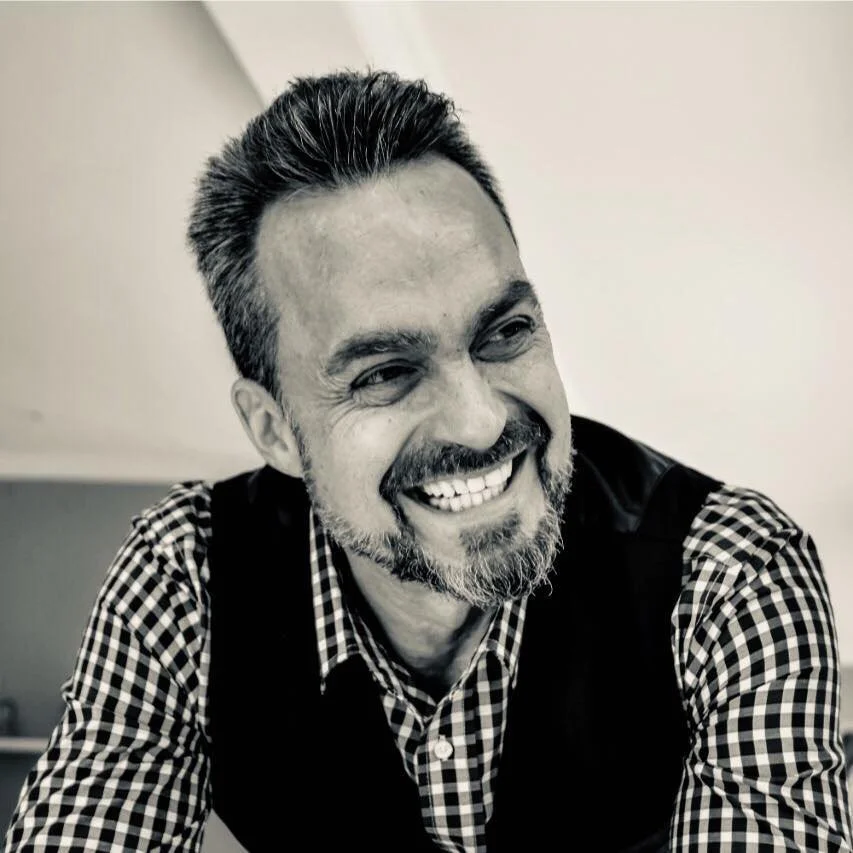 A smiling man with dark hair, a beard, wearing a checkered shirt and vest, in black and white photograph.