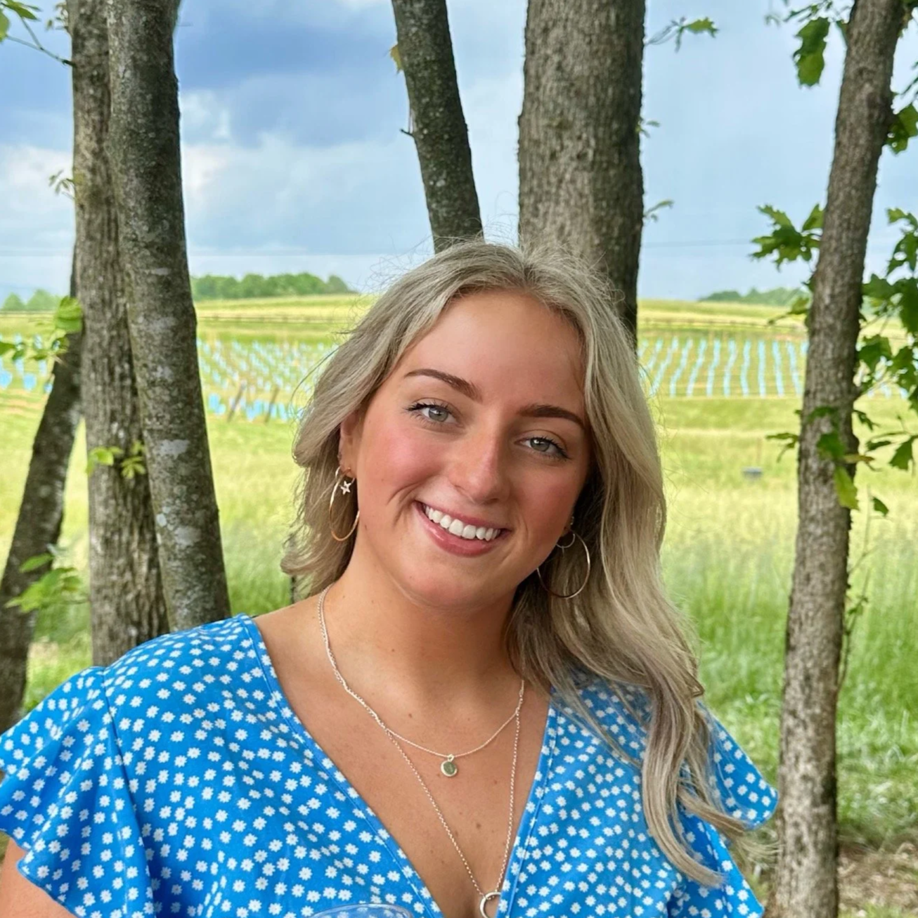 A smiling woman with blonde hair, wearing a blue and white polka dot dress, standing outdoors among trees with a field and a body of water in the background.