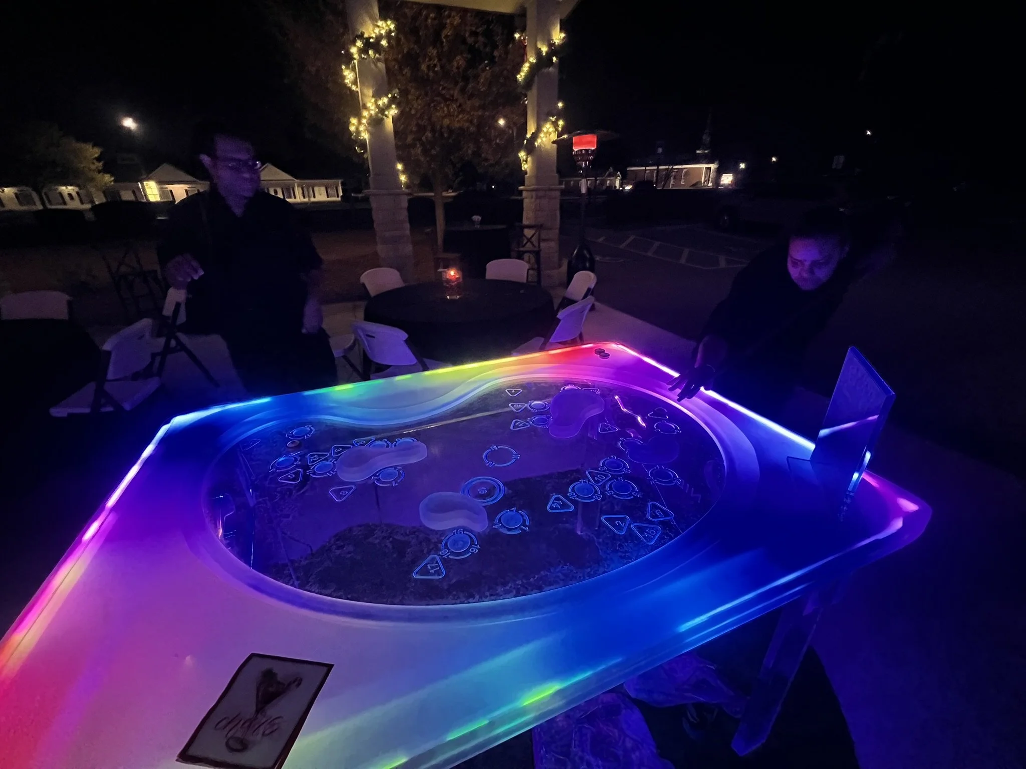A dark outdoor setting with a group of people gathered around a table illuminated by rainbow-colored neon lights, with trees decorated with string lights and a bright moon in the night sky.