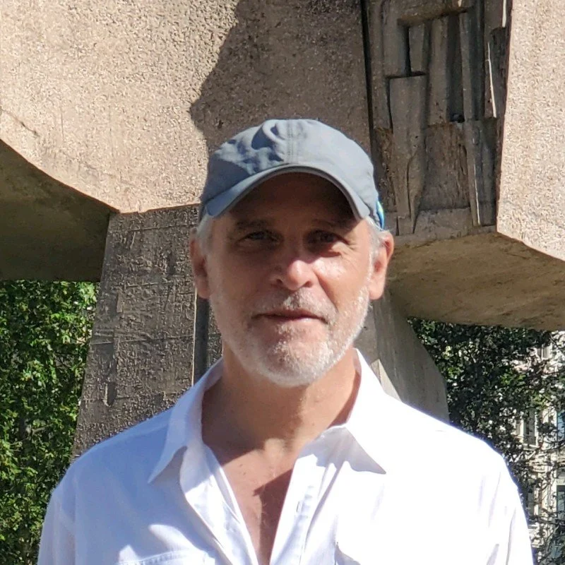 A middle-aged man with a gray beard and mustache wearing a light gray cap and a white collared shirt, standing outdoors in front of a concrete structure with trees in the background.