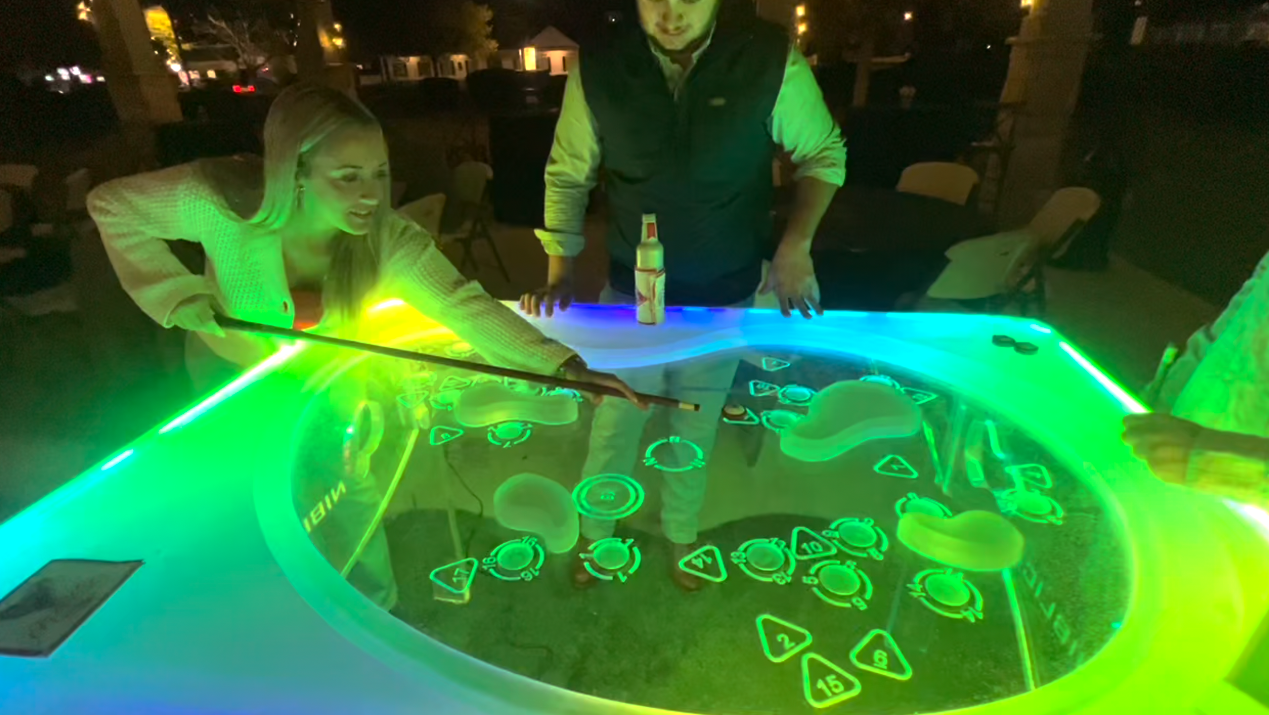 Two people are playing an illuminated electronic game at a table outdoors at night, with colorful glowing controls and a woman with long blonde hair holding a stick.