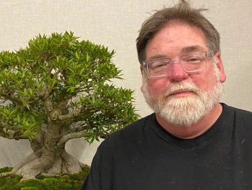 A man with glasses and a beard standing next to a large bonsai tree indoors.