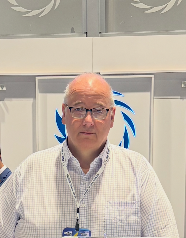 A middle-aged man wearing glasses and a white checkered shirt with a conference badge around his neck, standing in front of a backdrop with a blue and white logo.