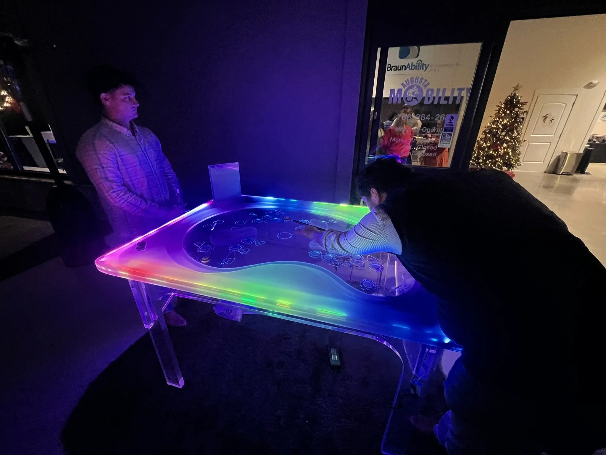 People playing air hockey with multicolored LED lights in a dark recreational room, decorated with a lit Christmas tree.
