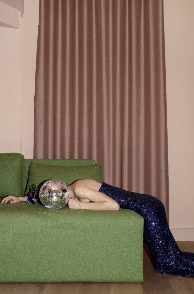 A person lying face down on a green sofa, wearing a black sequined dress, with a disco ball over their face, and a green glove on their left hand, in a room with pinkish-beige walls, a large pink curtain, and wooden floor.