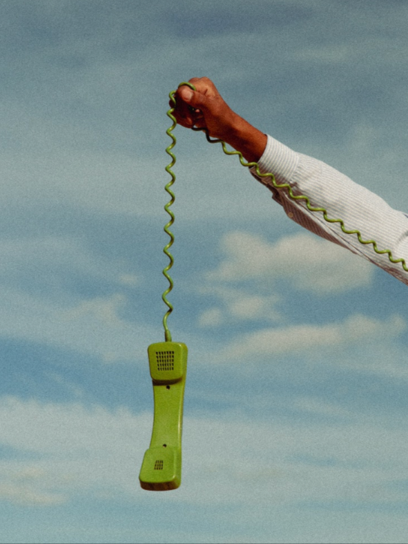 A person holding a green corded telephone receiver outdoors against a cloudy sky background.
