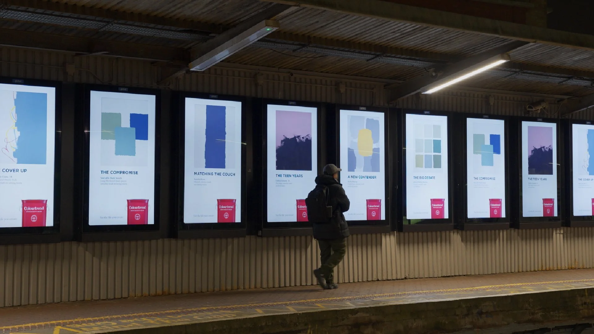 A person standing at a bus stop or train station, looking at digital screens displaying various colorful book covers and titles in an industrial setting with a corrugated metal wall and ceiling lights.