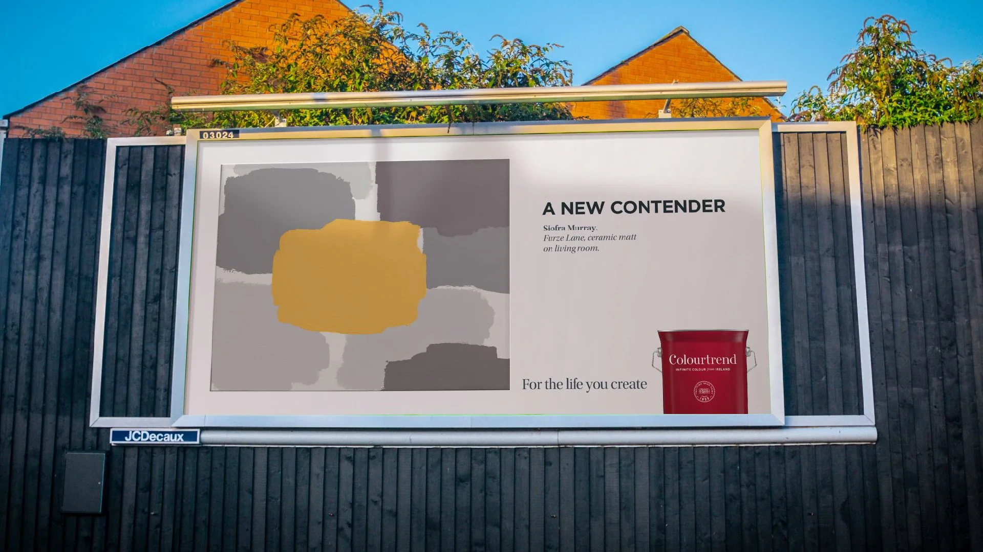Outdoor billboard advertising paint color 'A New Contender' by Colourtrend, featuring a color swatch and promotional text, mounted against a black wooden fence with a house and trees in the background.