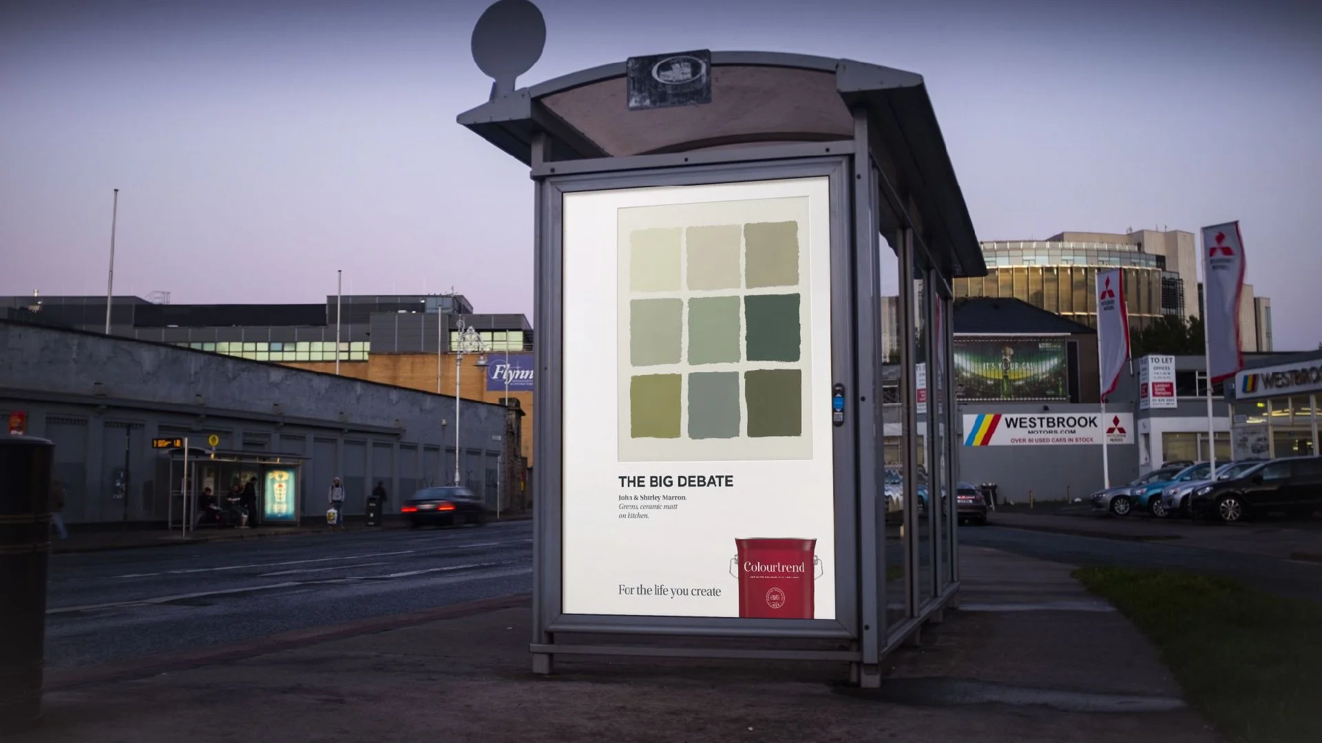 A bus stop advertising panel with a color paint palette and a red paint can labeled 'Colourtrend,' situated on a city street during dusk with cars and buildings in the background.