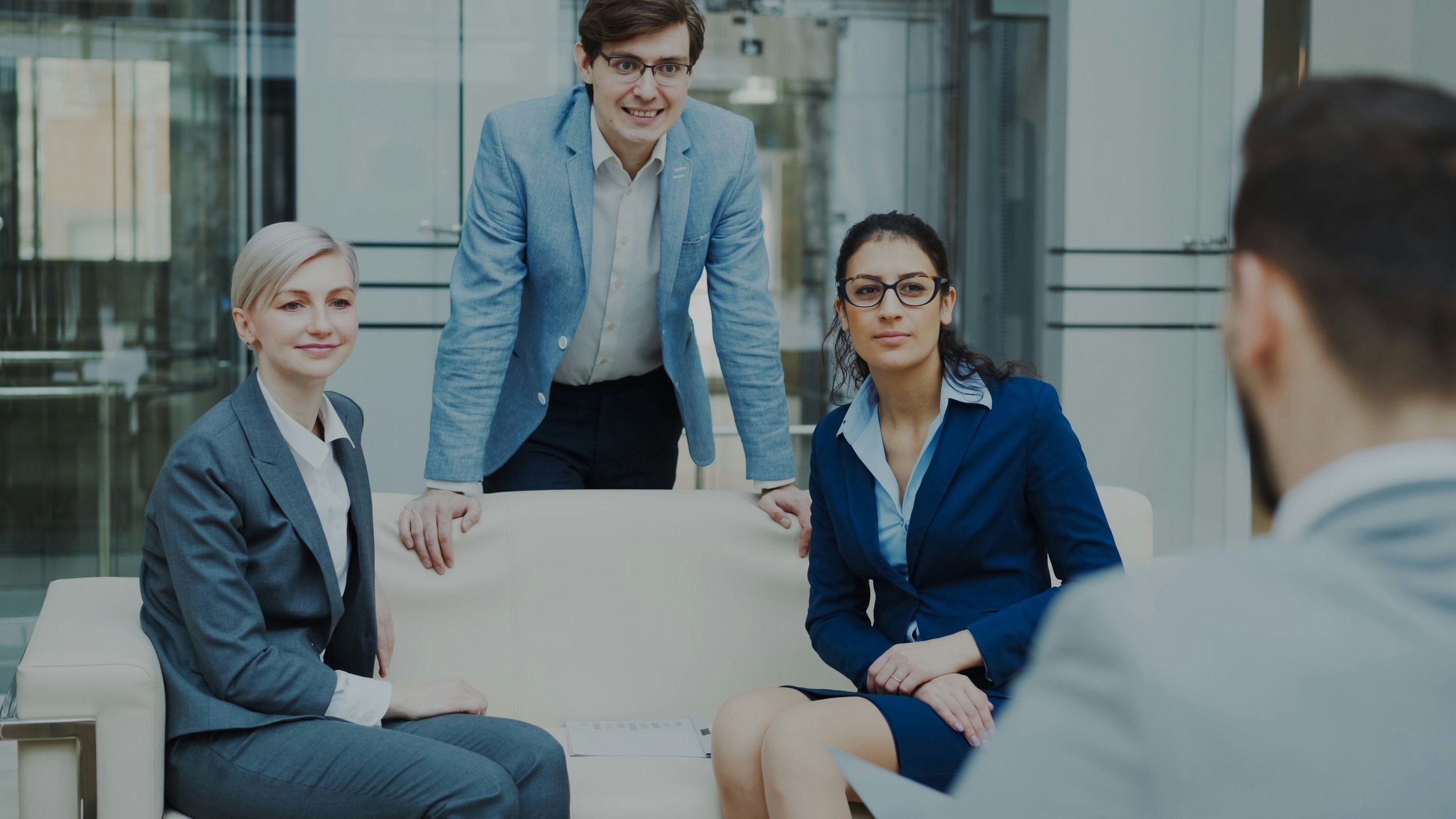 Quatre professionnels en réunion dans un espace de bureau moderne, trois femmes assises et un homme debout, discutant sérieusement.