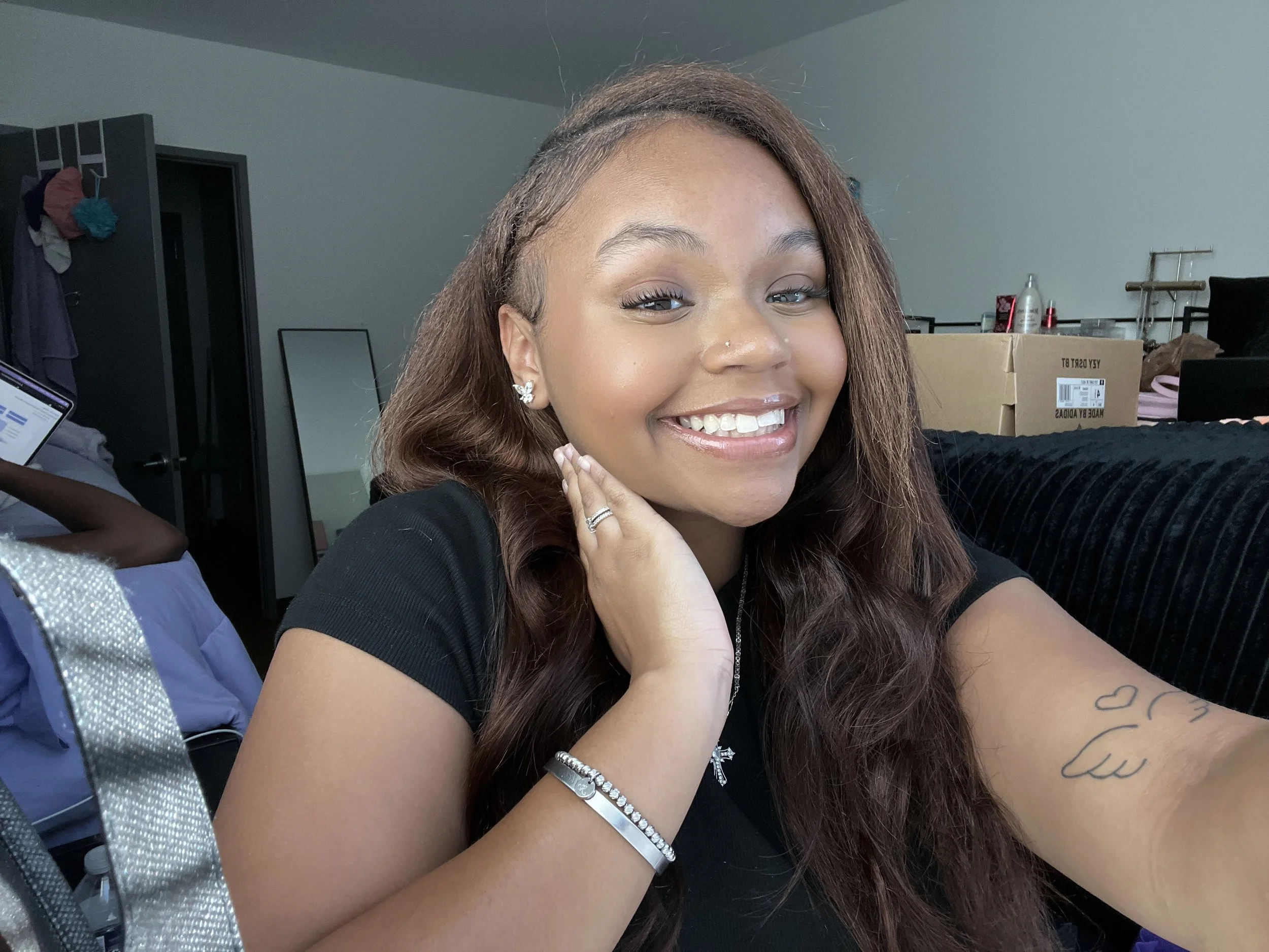 Smiling woman with long hair, wearing a black shirt, jewelry, and earrings, taking a selfie in a casual room.
