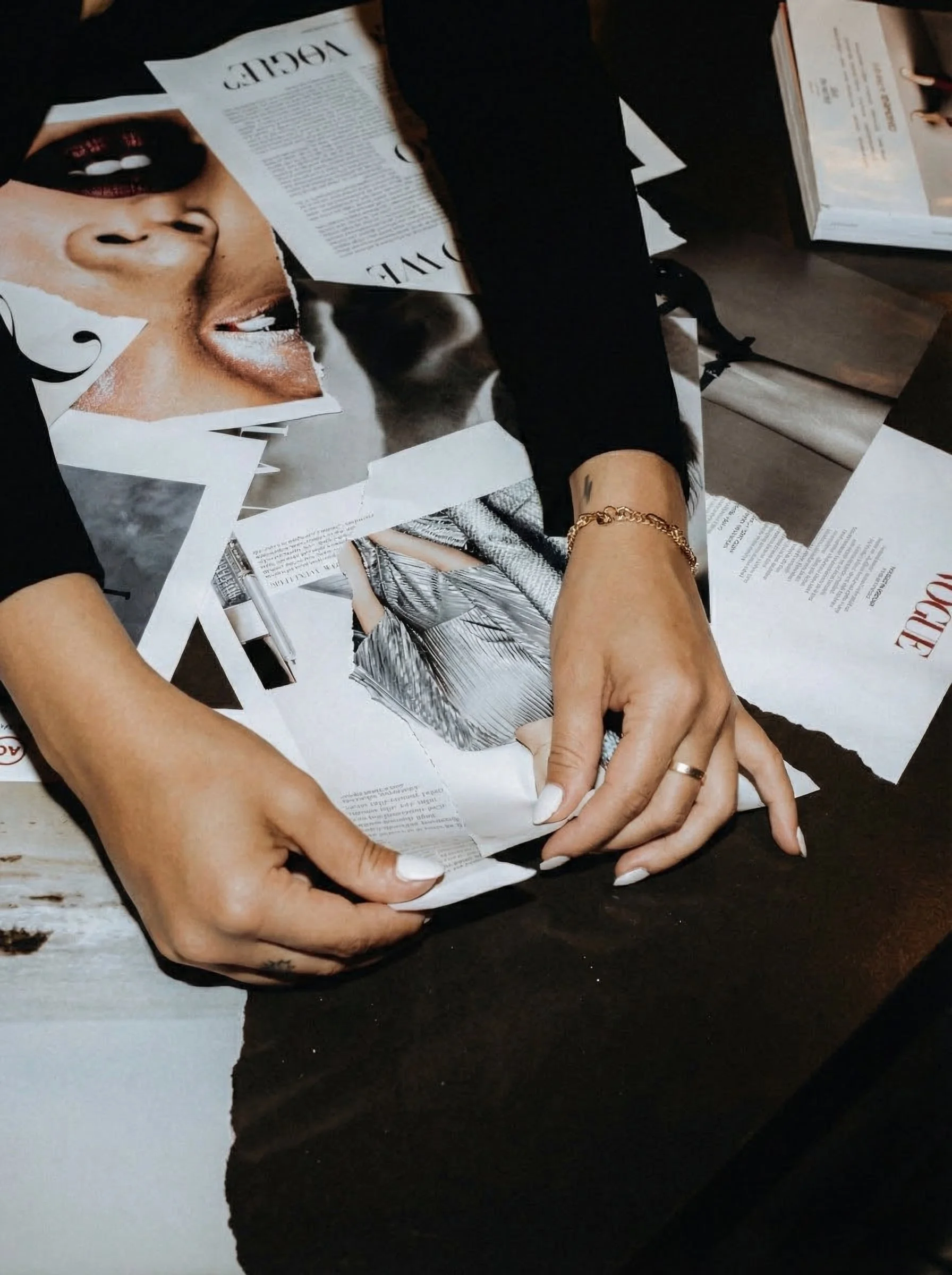 A person is folding torn magazine pages on a dark table, with fashion and beauty images visible.