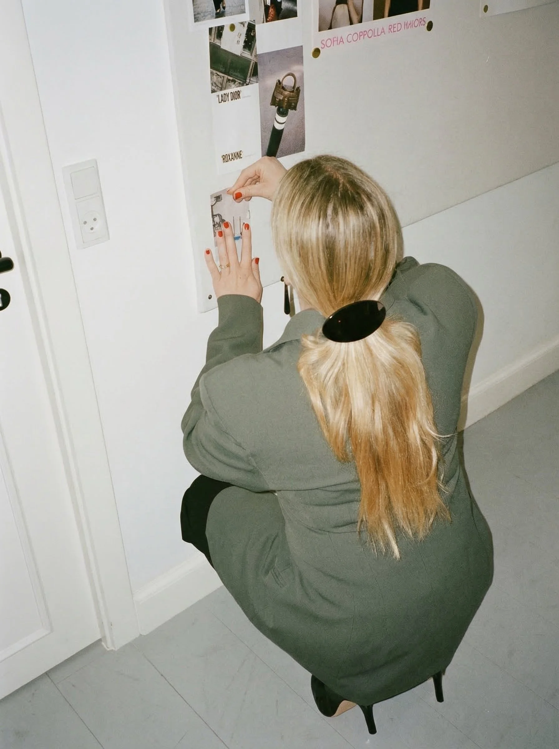 A woman with blonde hair and a black hair clip is squatting and posting something on a white wall covered with photos and notes, including one labeled 'Lady Dior' and another 'Sofia Coppola Red Mirrors.'